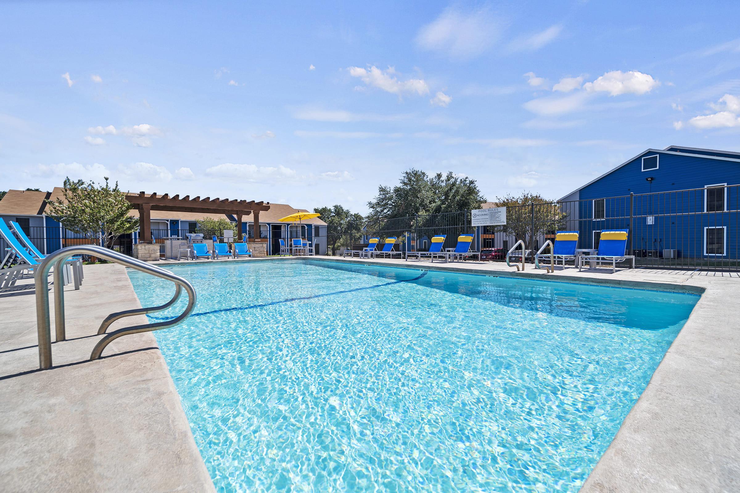 A bright blue swimming pool surrounded by lounge chairs and yellow umbrellas. In the background, there are buildings with a blue exterior and green trees. The pool is clear and reflects the sky, creating a serene atmosphere for relaxation.