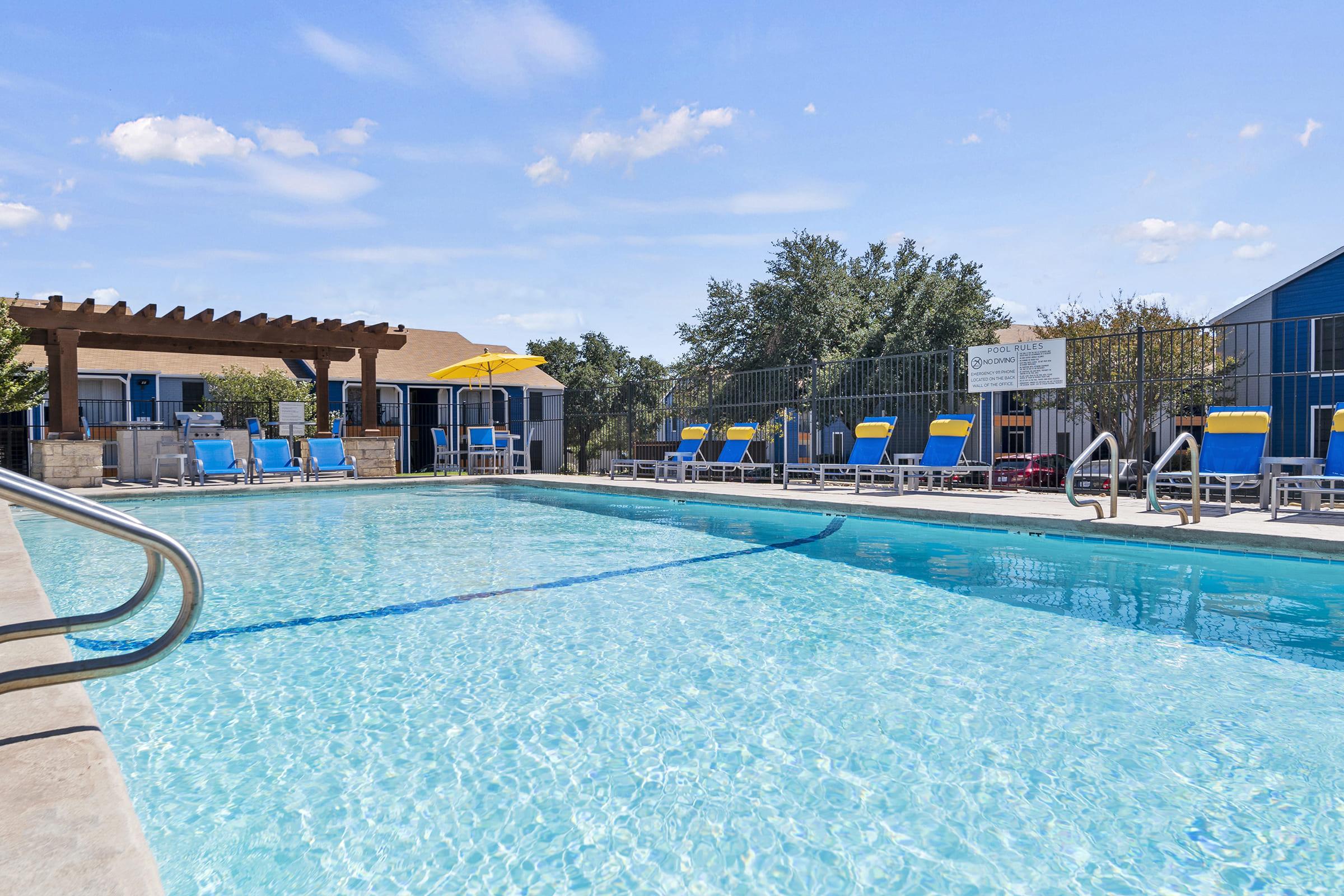 A sparkling blue swimming pool surrounded by lounge chairs under a sunny sky. There is a shaded pergola on one side, and a yellow umbrella provides additional shade. Nearby, a few residential buildings are visible, adding to the inviting atmosphere of the outdoor space.