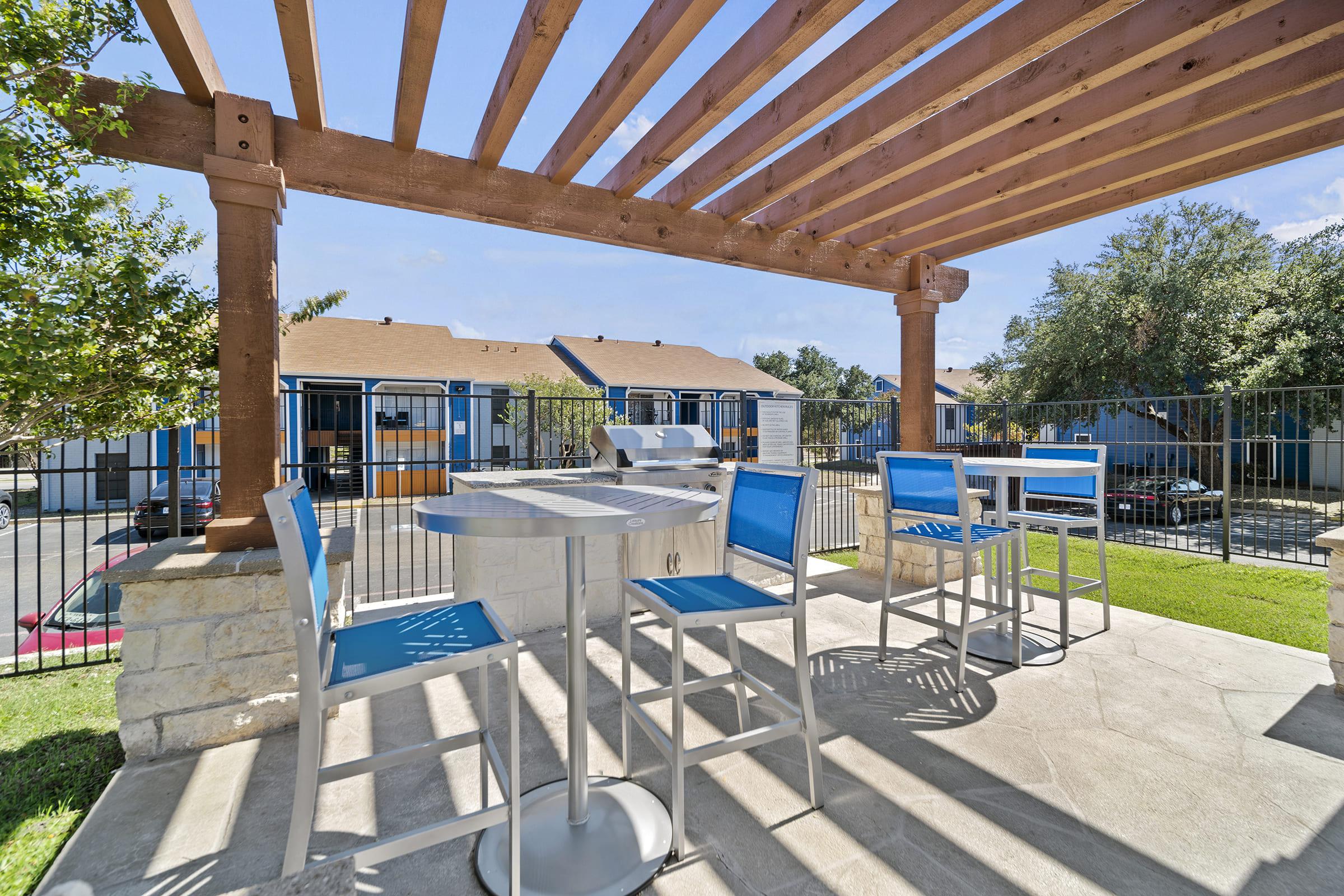 A shaded outdoor patio area featuring a modern table and chairs, with a grill nearby. The setting includes a backdrop of multiple buildings and greenery, creating a relaxed atmosphere ideal for social gatherings or dining al fresco.