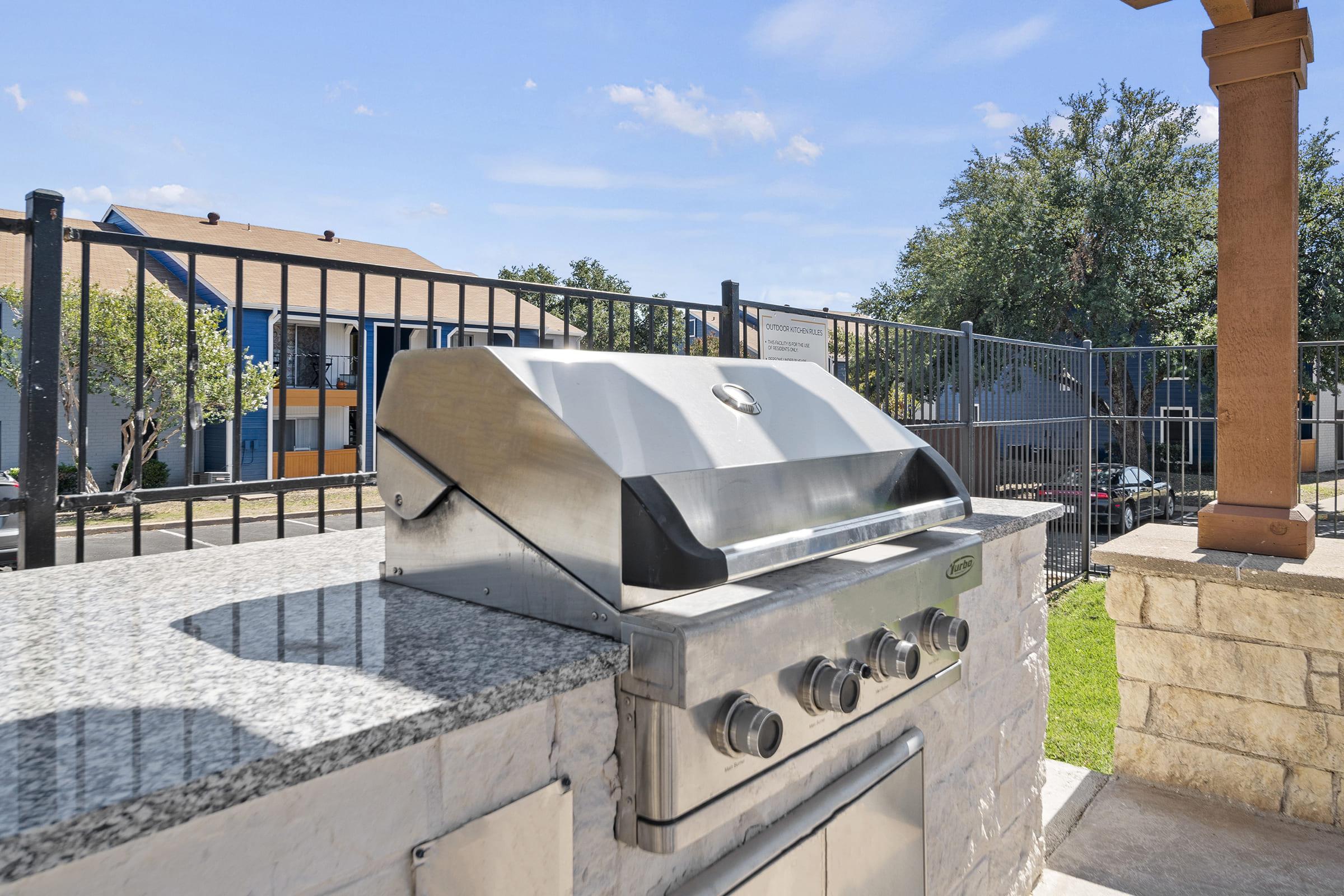 Stainless steel grill on a stone countertop with a clear blue sky and buildings in the background. The grill has four control knobs and is situated in an outdoor area, surrounded by a black fence and green grass.