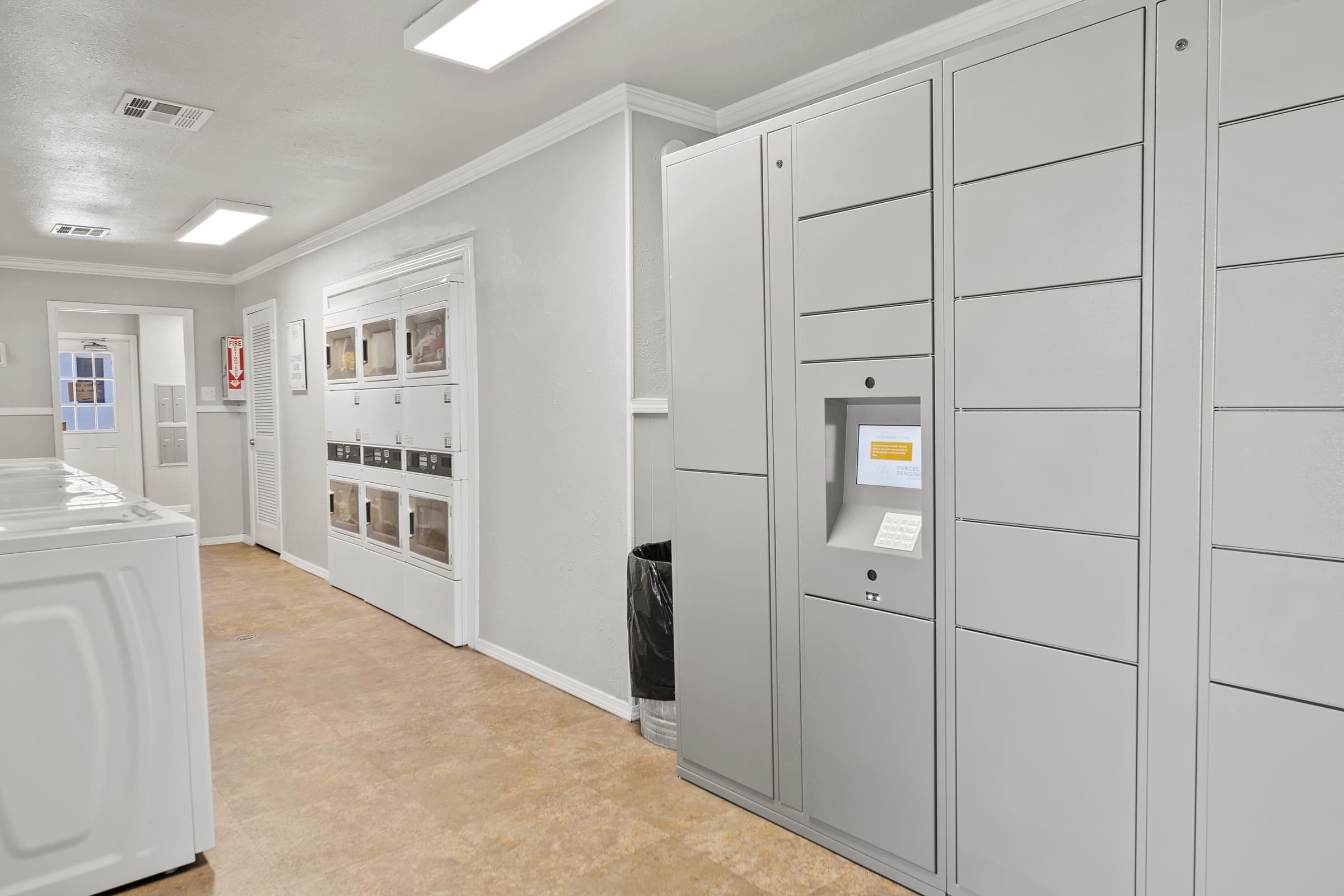 A modern laundry room featuring white machines on the left, rows of storage lockers on the right, and a parcel locker with a digital interface. The walls are painted light gray, and there are overhead lights illuminating the space. A trash can is positioned near the lockers.