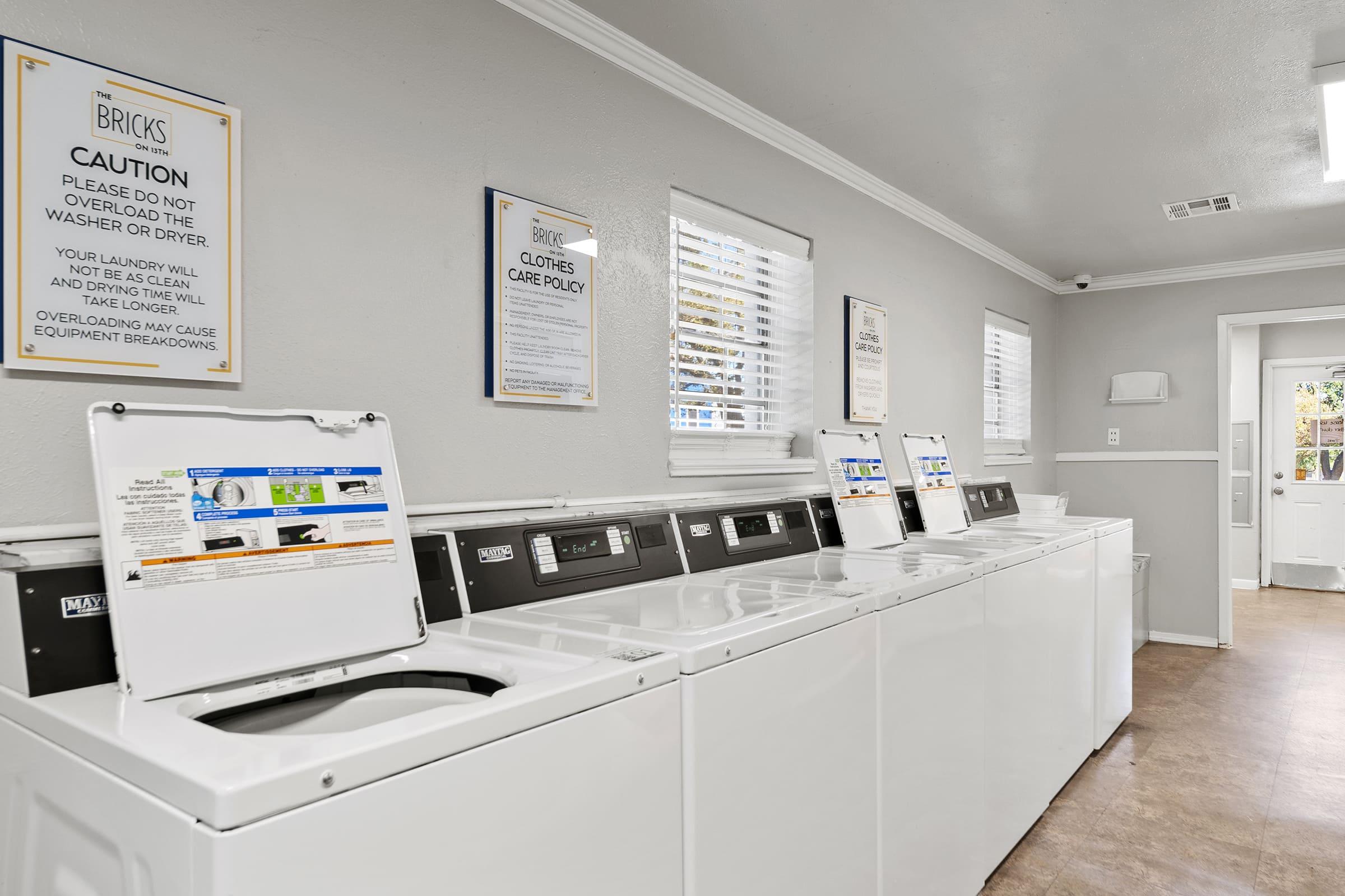 A clean laundry room featuring several stacked washing machines and dryers. The walls are painted light gray, and there are framed signs providing caution and care instructions. Natural light enters through windows, and the overall space is neat and organized.