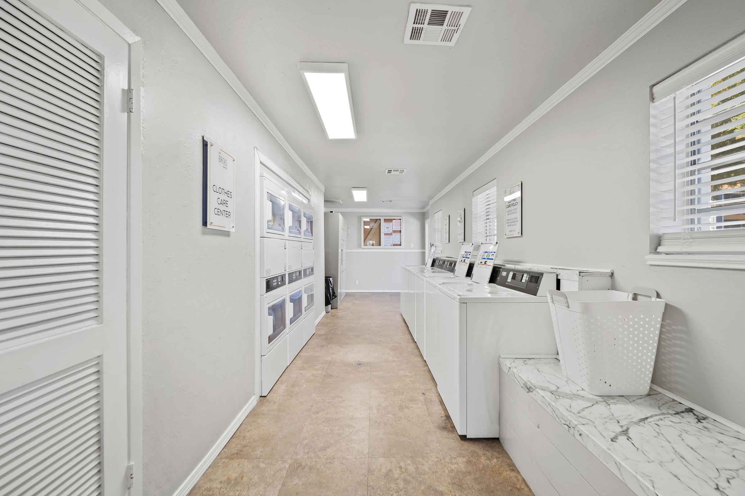 A clean, well-lit laundry room featuring white walls and tiled flooring. Several washing machines and dryers are lined along one side, with a large white laundry basket on a marble-topped seating area. Windows provide natural light, and bulletin boards with information are visible on the walls.