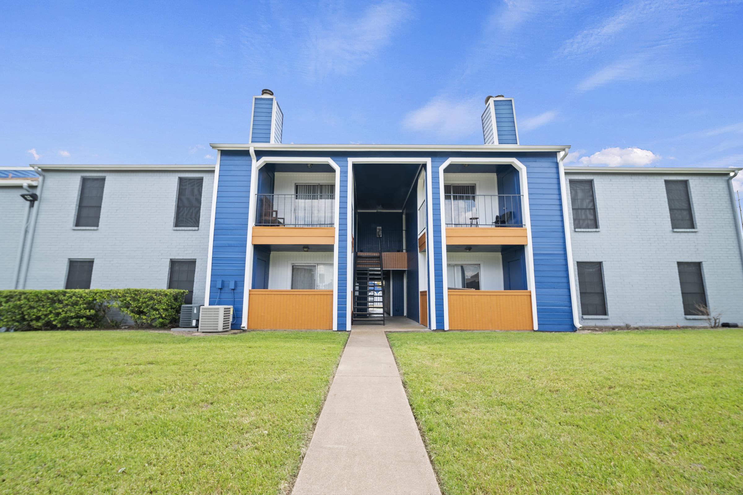 A two-story apartment building with blue and orange accents, featuring balconies on the upper floor. The building has a small walkway leading to the entrance and a grassy area in front. Large windows with screens are visible, and there are air conditioning units installed.