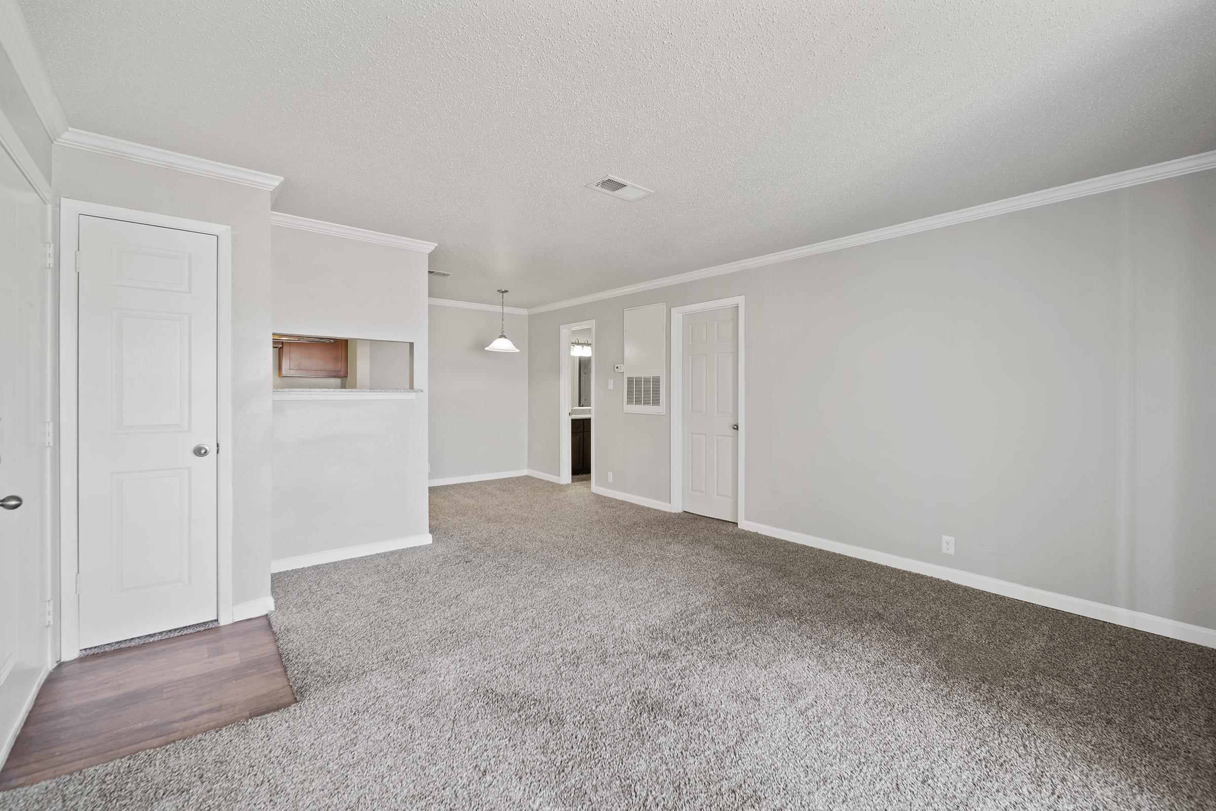 A spacious, well-lit living area featuring light gray walls and carpet, with a doorway leading to a bathroom and a small kitchen area visible in the background. The room has a pendant light fixture and a door on the right leading to another room.