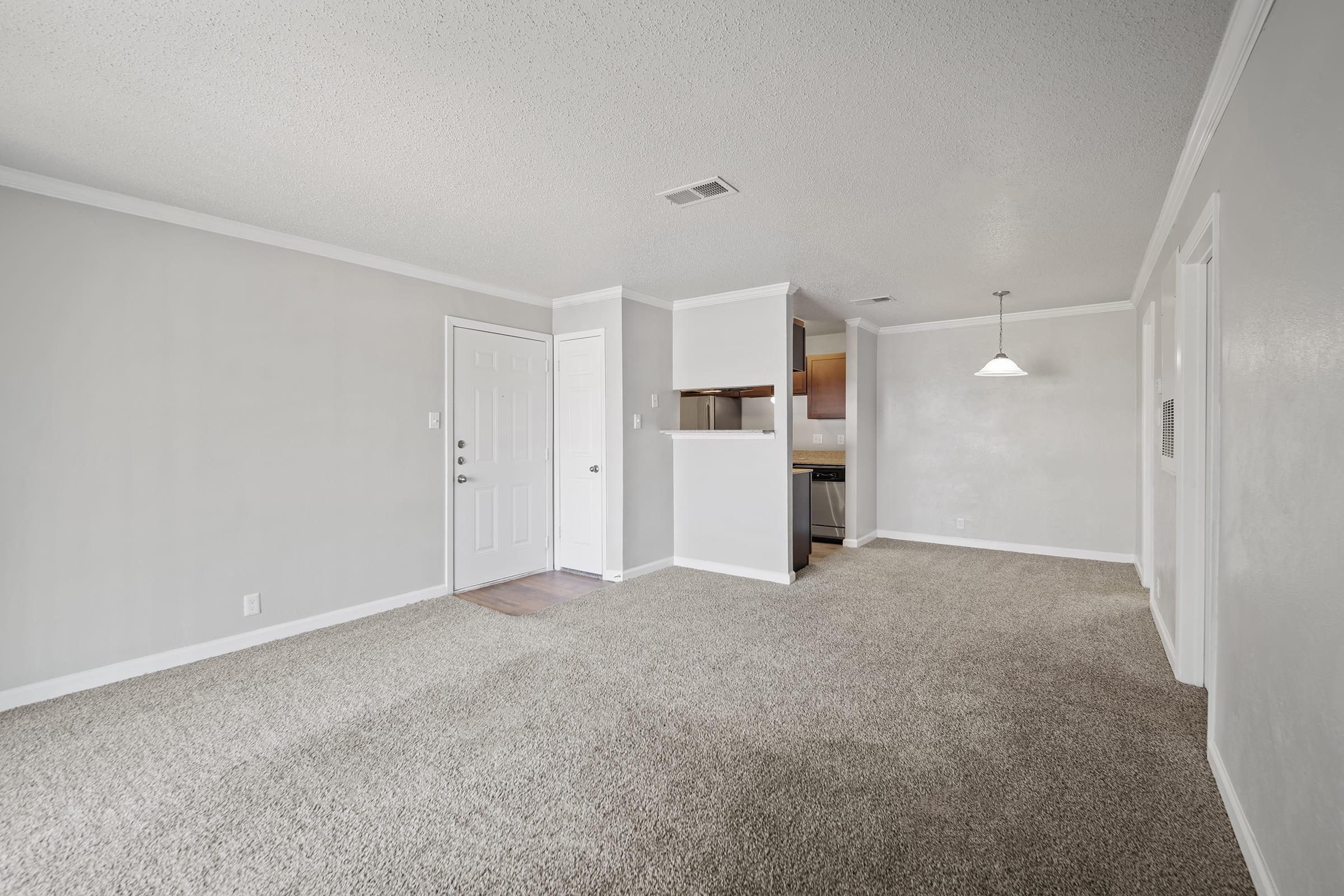 A spacious living area with light gray walls and cozy carpet flooring. A front door is visible, leading to the entrance. An open layout connects to a kitchen area with a small bar. A pendant light hangs from the ceiling, creating a warm ambiance. The space appears well-lit and inviting.