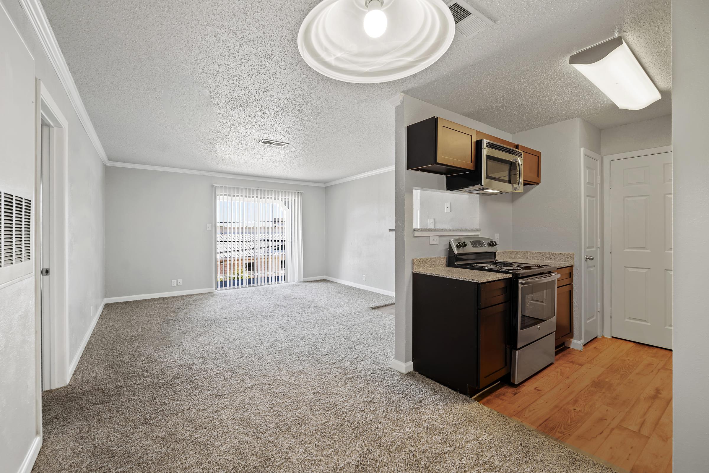 Bright, open living space featuring light-colored walls, plush carpet, and wood flooring. The kitchen is equipped with modern appliances, including a stainless steel stove and microwave, and a small breakfast bar. Natural light streams in through sliding glass doors, leading to an outdoor area.