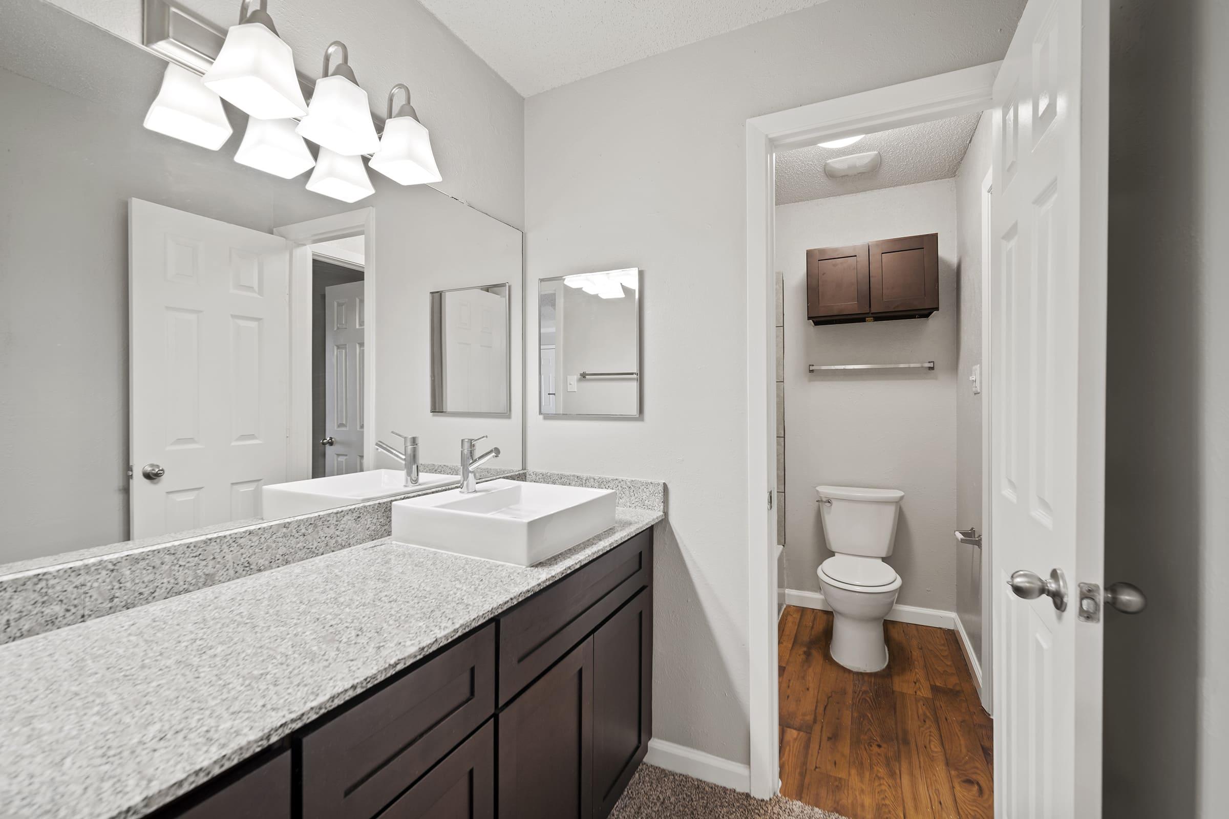 Modern bathroom featuring gray walls, a double sink with undermount white basins, and a granite countertop. Two mirrors are above the sinks, and there is a doorway leading to a separate toilet area with a wooden floor. Ample lighting from stylish light fixtures above the sinks.