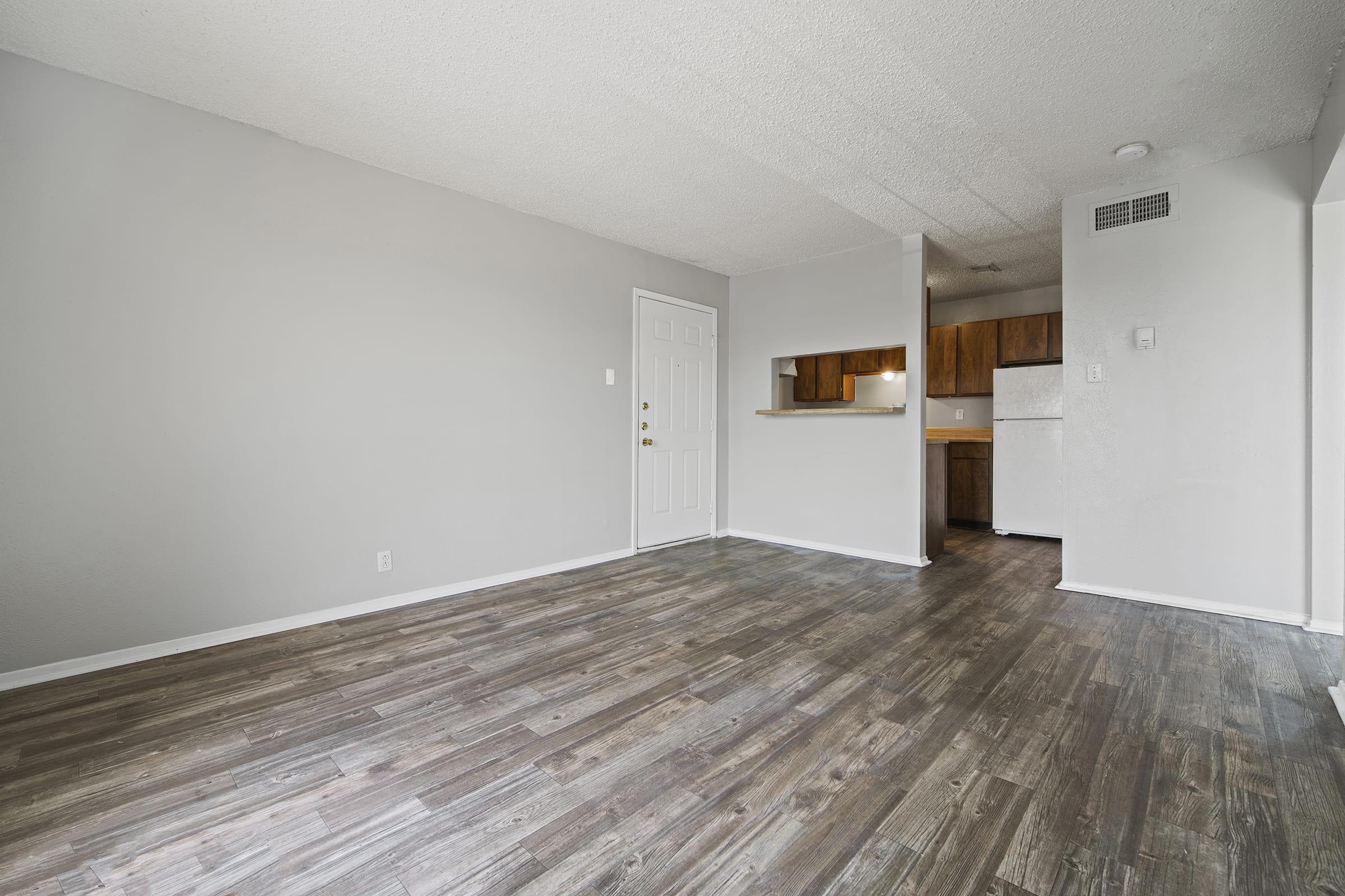 An empty room with gray walls and wooden laminate flooring. To the left, there's a front door. In the background, a small kitchen area with wooden cabinets and a refrigerator is visible. The space is well-lit with natural light, creating a clean and open atmosphere.