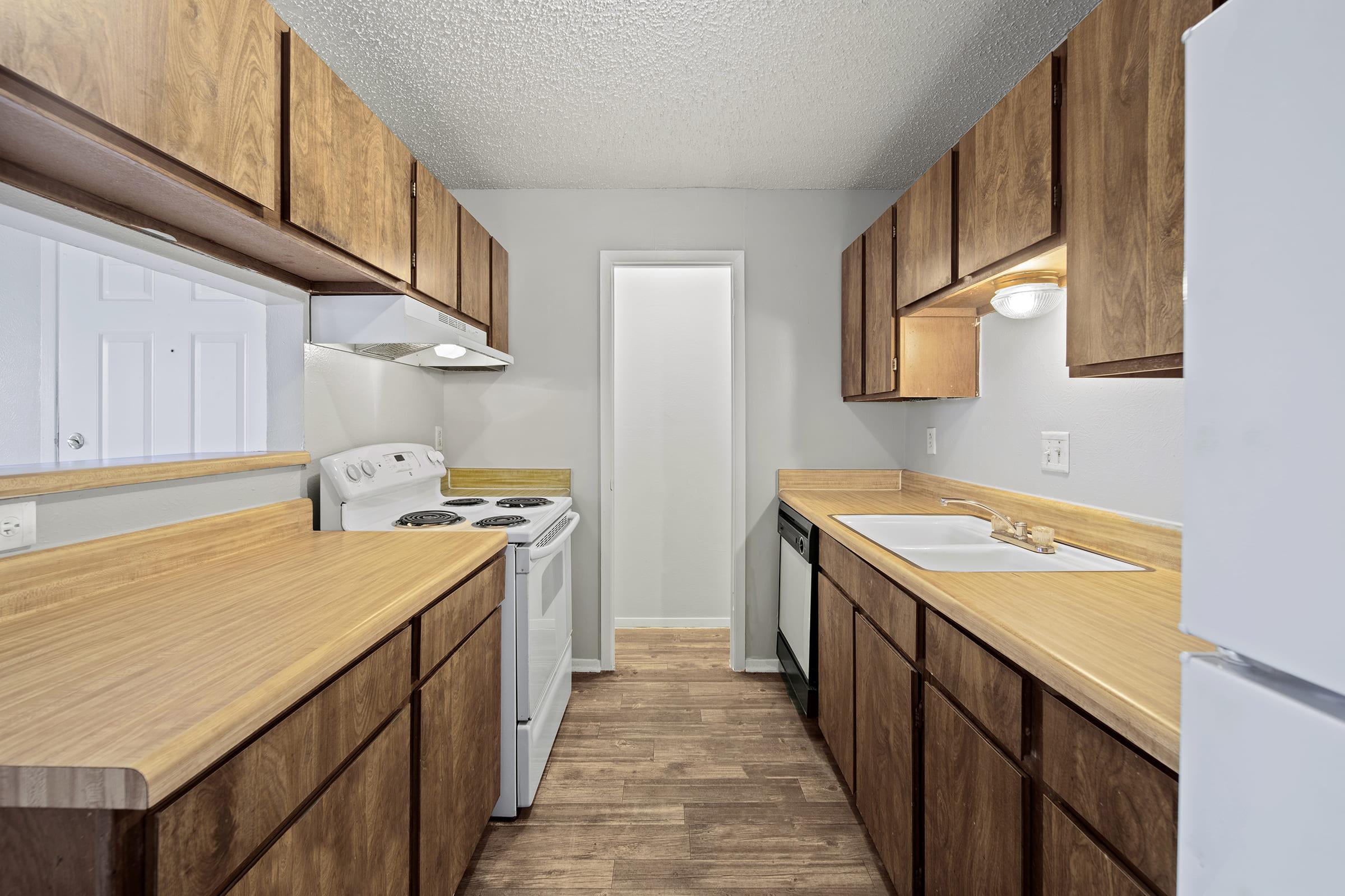 A well-lit, modern kitchen featuring wooden cabinets, a white stove, and a sink. The countertops are a light wood finish. A refrigerator is on the right, and a door leads to another room at the end of the kitchen. The floor has a wood-like appearance, enhancing the kitchen's warm aesthetics.