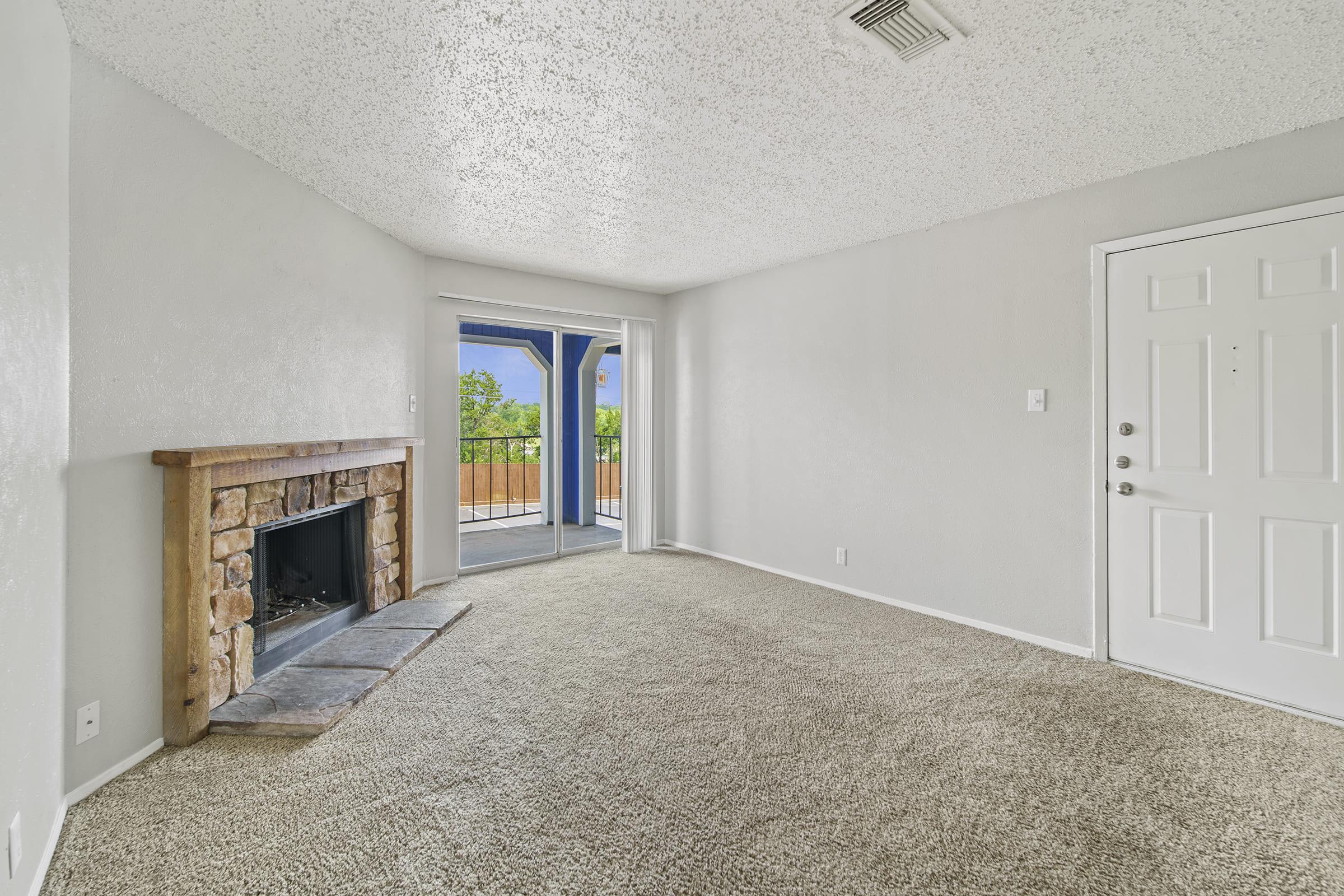 A living room with a cozy stone fireplace, light-colored textured walls, and plush carpeting. Large sliding glass doors lead to a patio area outside, allowing natural light to fill the space. The room features a simple white door and a ceiling textured with a speckled finish.