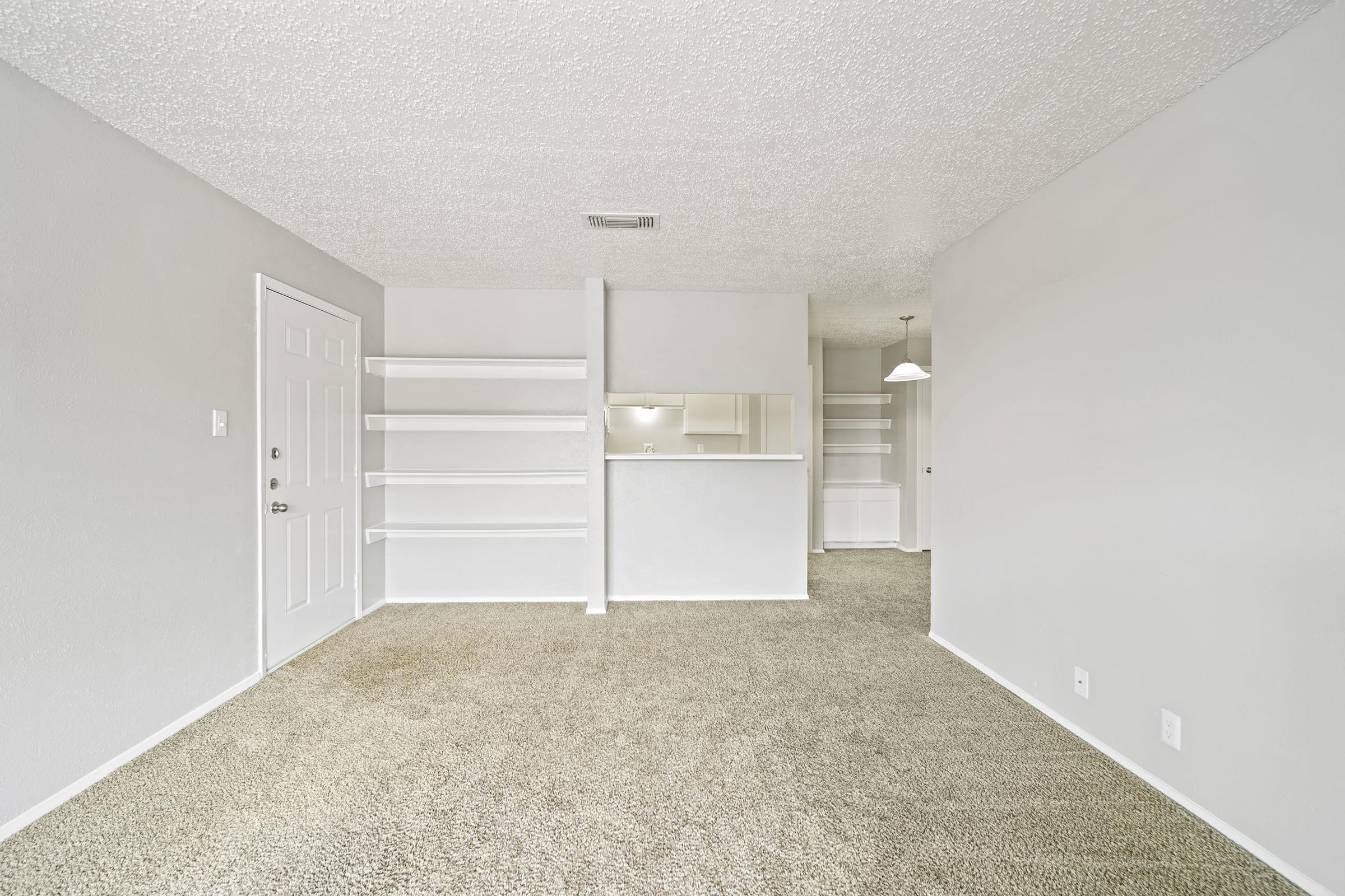 A spacious, empty living room featuring beige carpeting, white walls, and a door leading outside. There are built-in shelves on the left side and a small bar area facing the kitchen, with a light fixture hanging in the background. The room has a bright and airy feel.