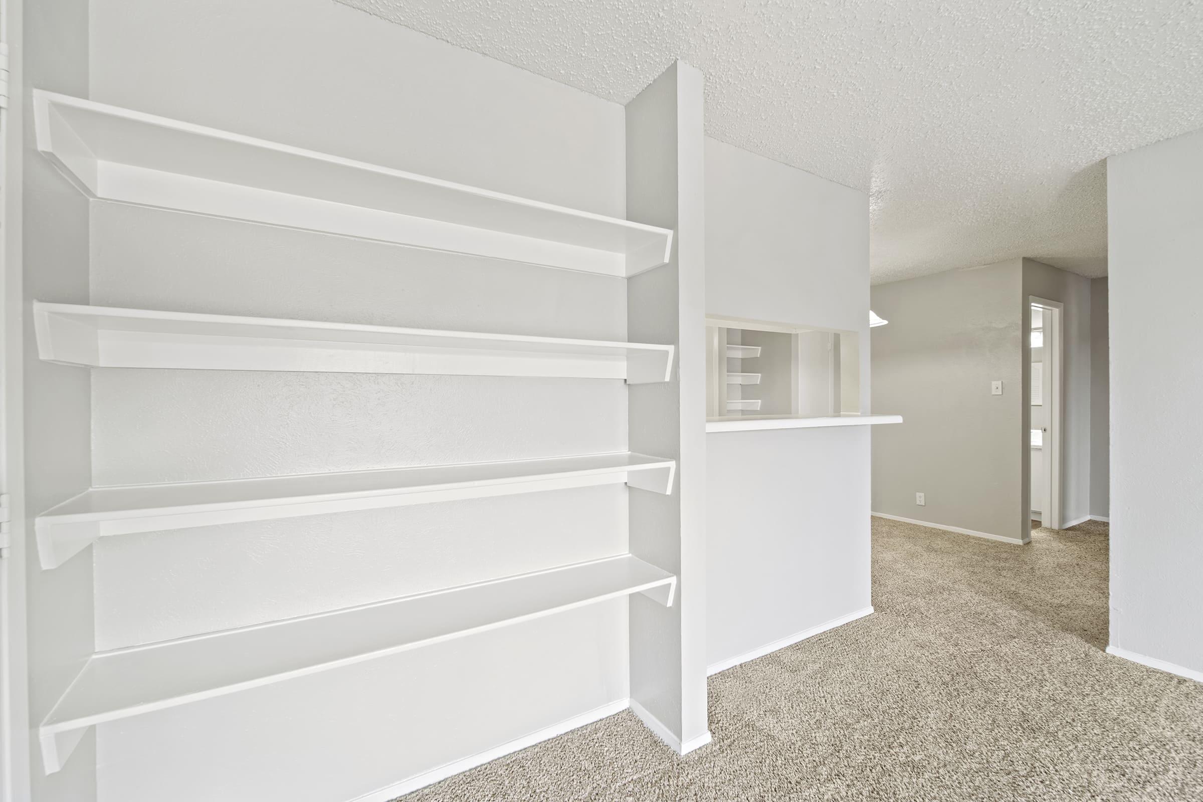A brightly lit room with light gray walls and beige carpet. On the left, there are three open white shelves against a wall, and in the background, a doorway leads to another part of the space. The overall atmosphere appears modern and well-maintained.