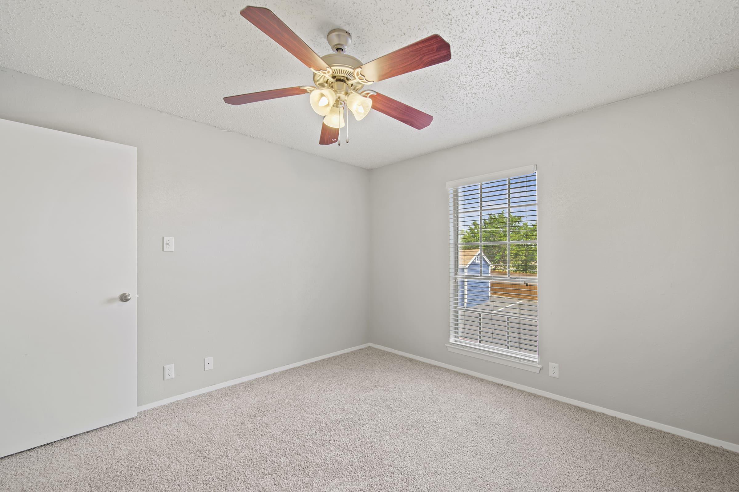 A well-lit bedroom featuring a ceiling fan with wooden blades, light-colored walls, and a plush carpeted floor. A window with blinds lets in natural light, revealing views of nearby rooftops outside. The room includes a white door and minimal decor, creating a clean and spacious appearance.