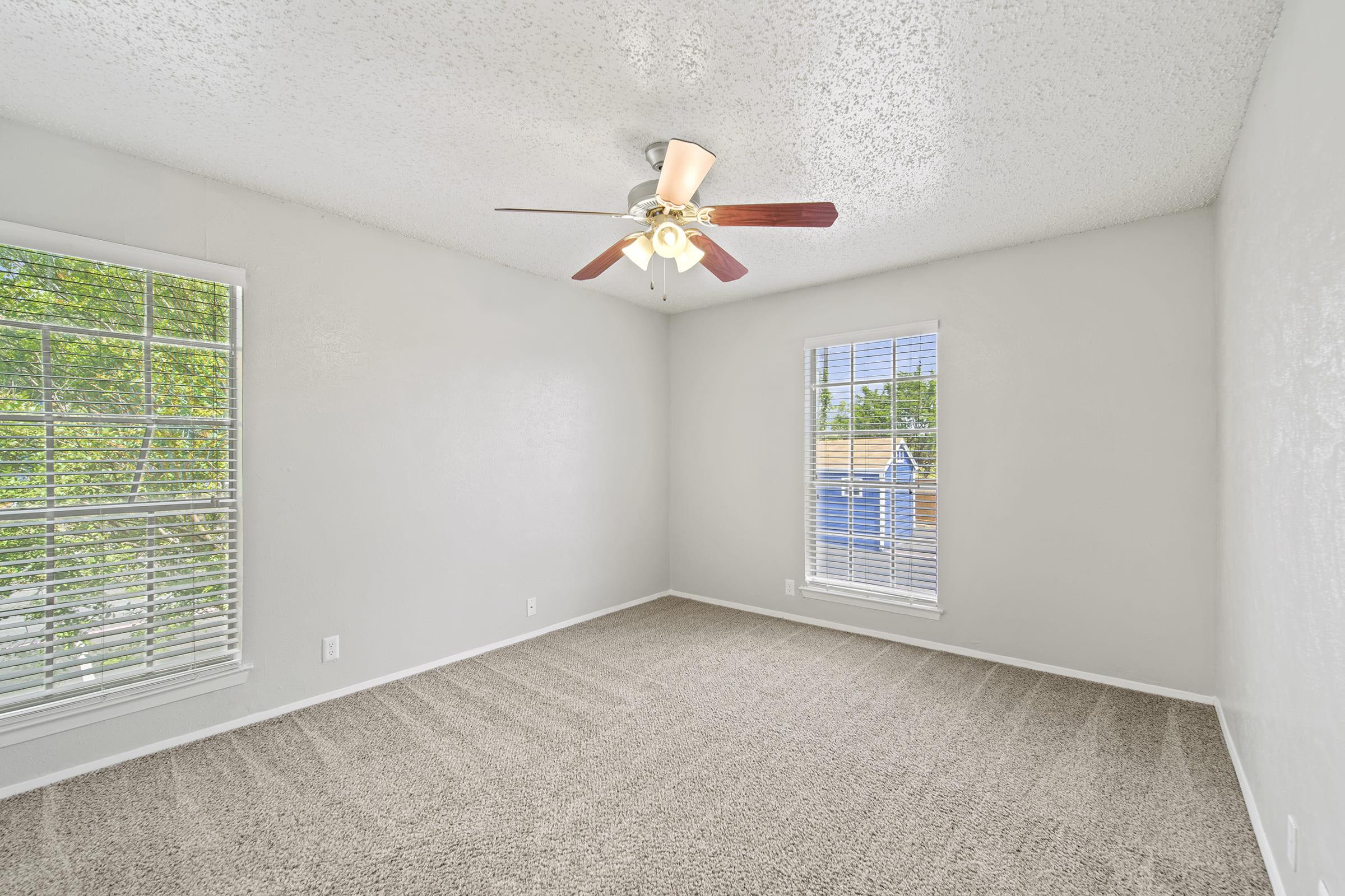 A vacant room with light gray walls and a ceiling fan, featuring two windows with white blinds. The floor is covered in soft beige carpet, and natural light floods the space, creating a bright and airy atmosphere.