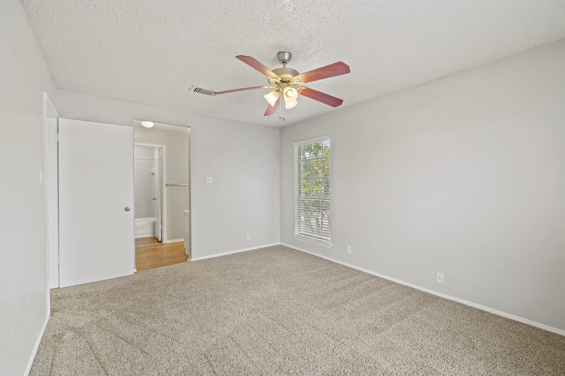 A spacious, empty room with beige carpet, a ceiling fan with red blades, and light gray walls. A doorway leads to another room, and a window provides natural light. The overall atmosphere is bright and airy, ideal for decorating.