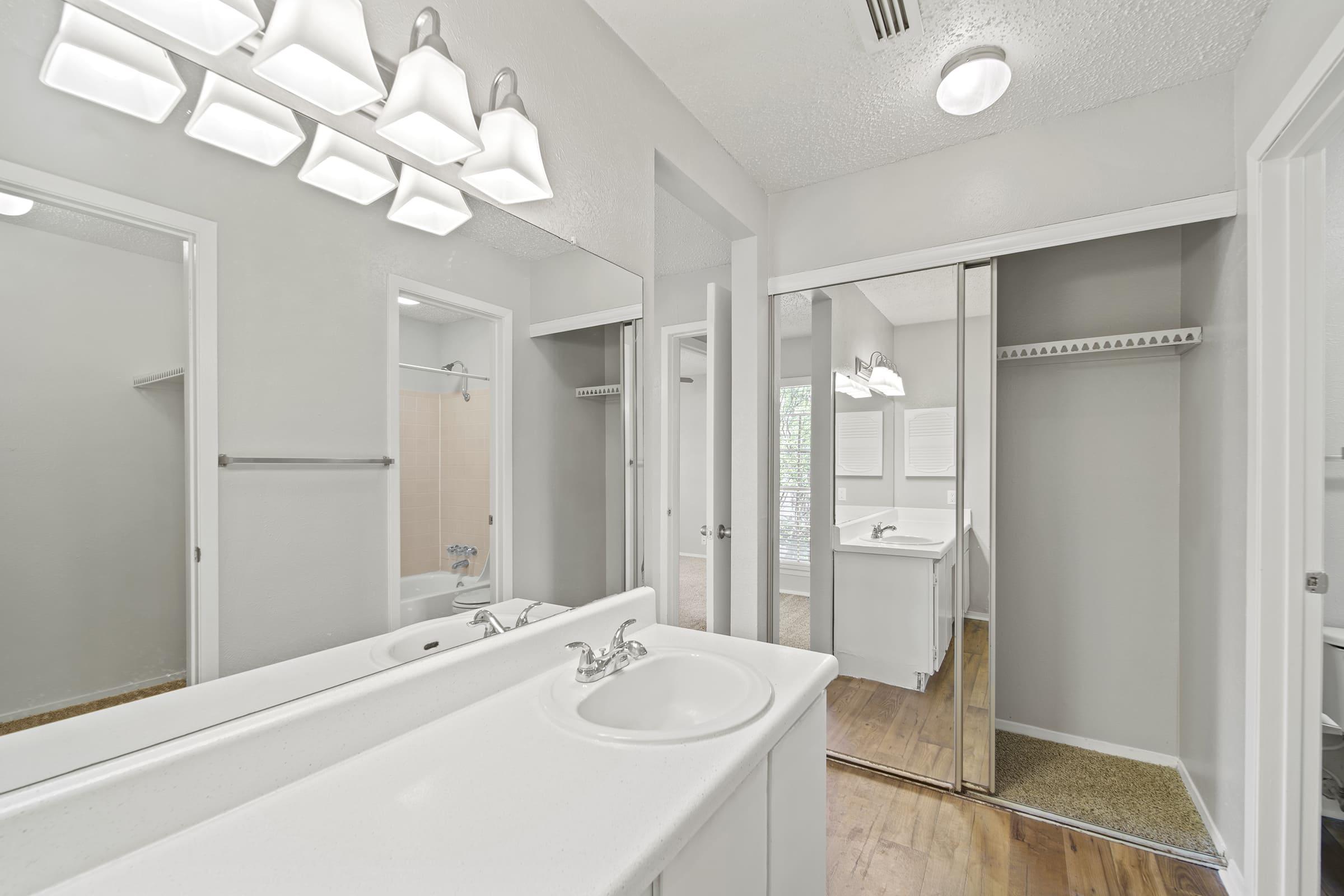 A clean and modern bathroom featuring a double vanity with a white countertop, ample mirrors, and a bright light fixture. The space includes light gray walls, a walk-in closet with sliding doors, and a shower area visible in the background. Hardwood flooring adds warmth to the design.