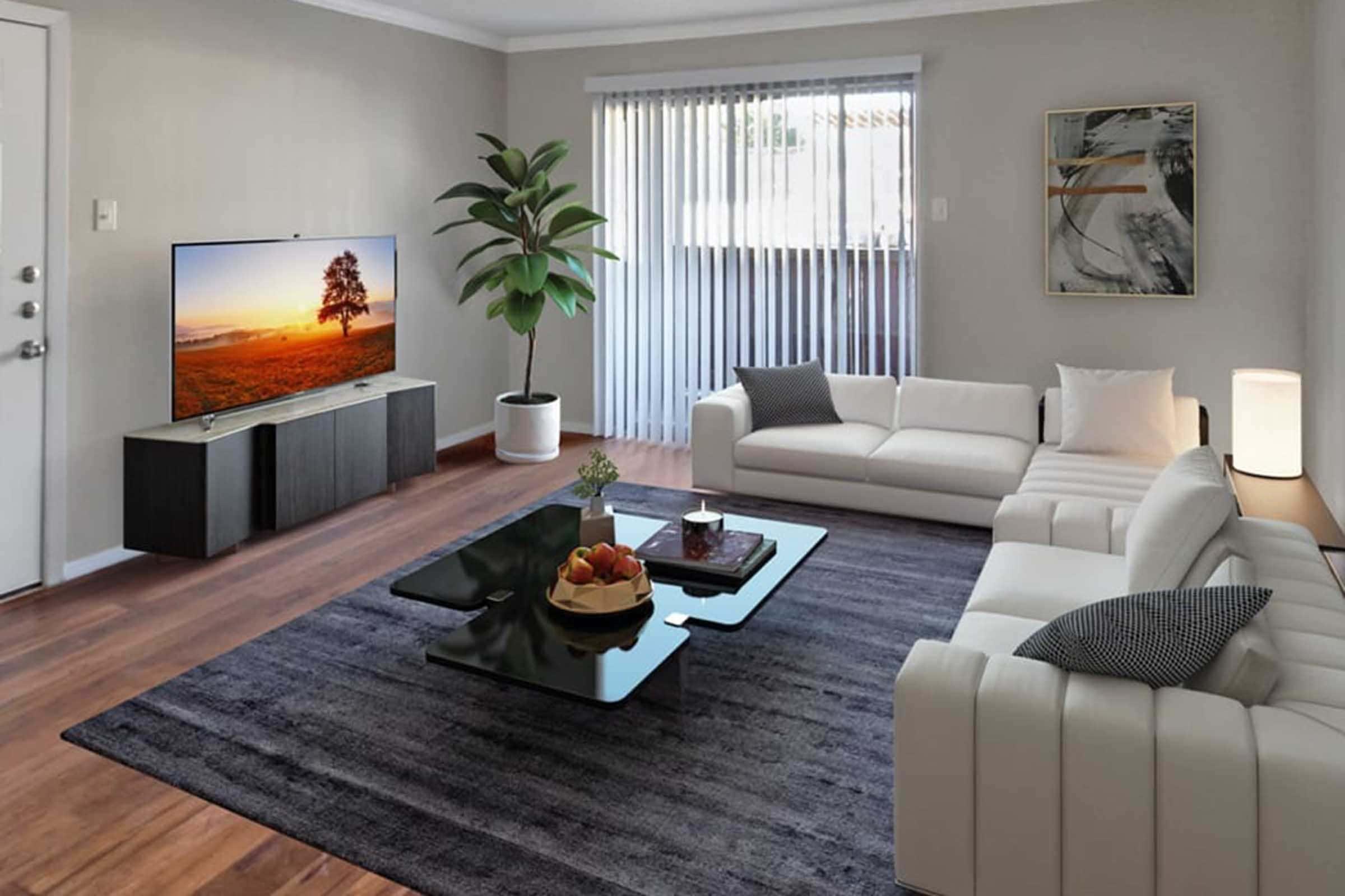 A modern living room featuring two white sofas, a glass coffee table with a fruit bowl, and a large TV on a black cabinet. A tall plant in a white pot adds greenery, while natural light filters through sheer curtains. The wooden flooring complements the contemporary design.