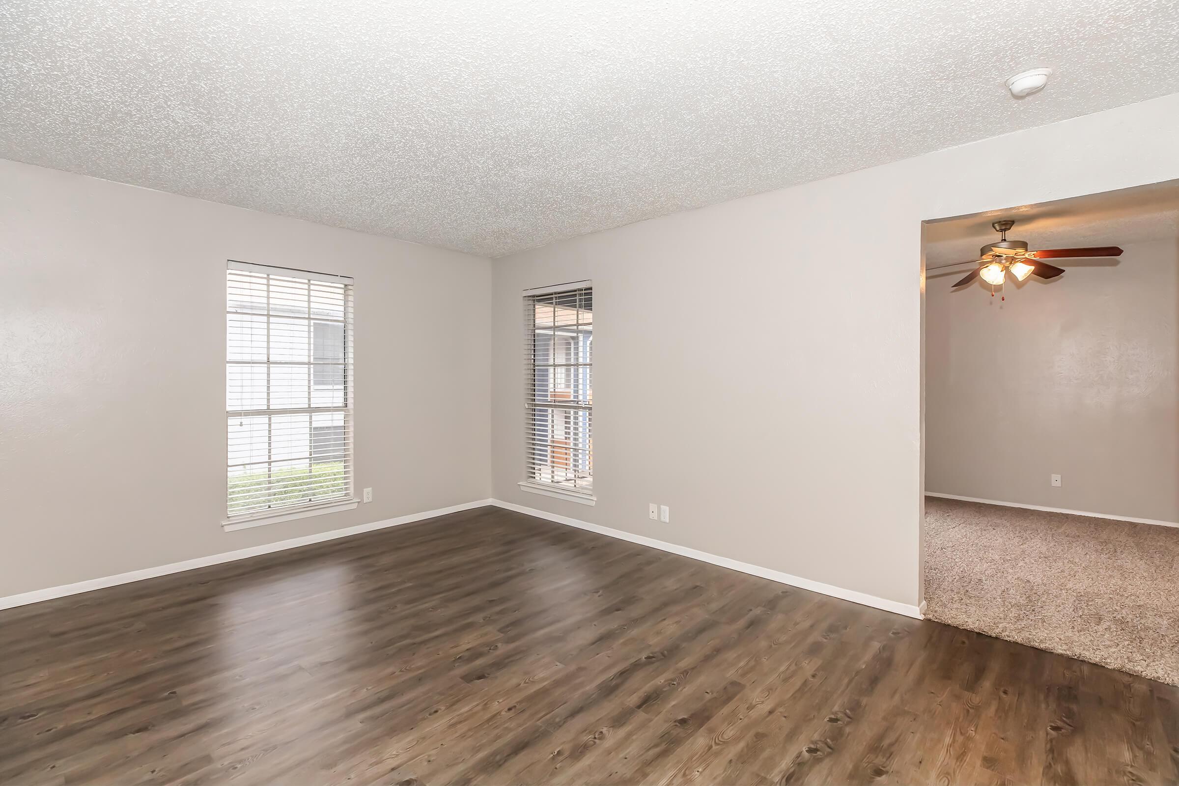 Spacious living area with light-colored walls and large windows, creating a bright atmosphere. The floor is covered with dark wood-like laminate. A doorway leads to a carpeted room with a ceiling fan, enhancing the room's charm. Ideal for a cozy and welcoming space.