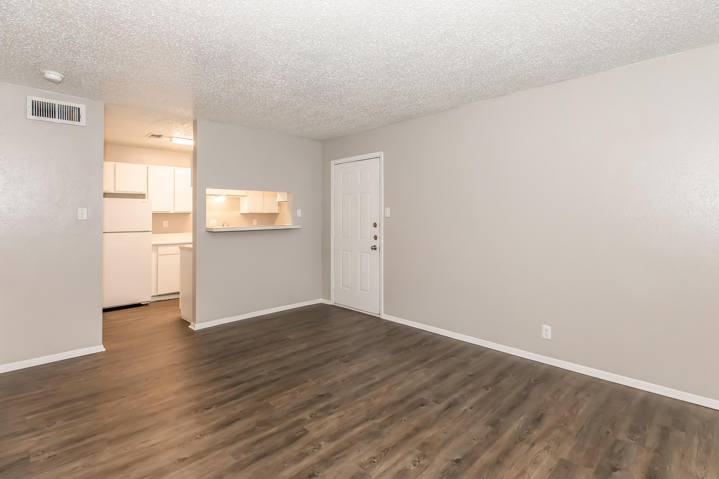 Spacious living area with light-colored walls and modern laminate flooring. A doorway leads to the entrance, while a kitchen area is visible in the background through an open pass-through. Natural light may be present, enhancing the inviting atmosphere of the space.