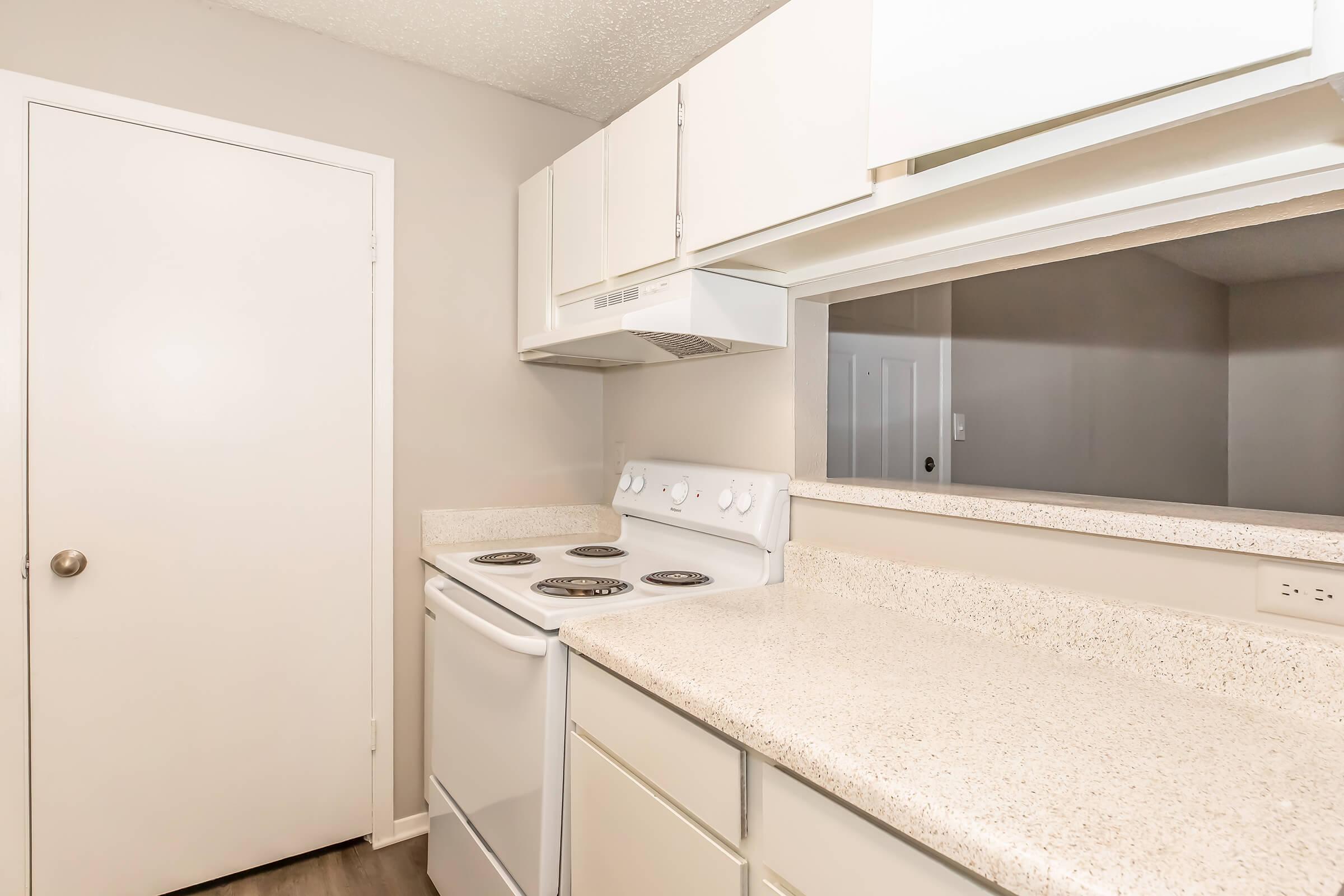 A modern kitchen featuring a white stove and oven, beige countertops, and white cabinetry. There's a door on the left and an open space leading to another room in the background, with neutral-colored walls and flooring.