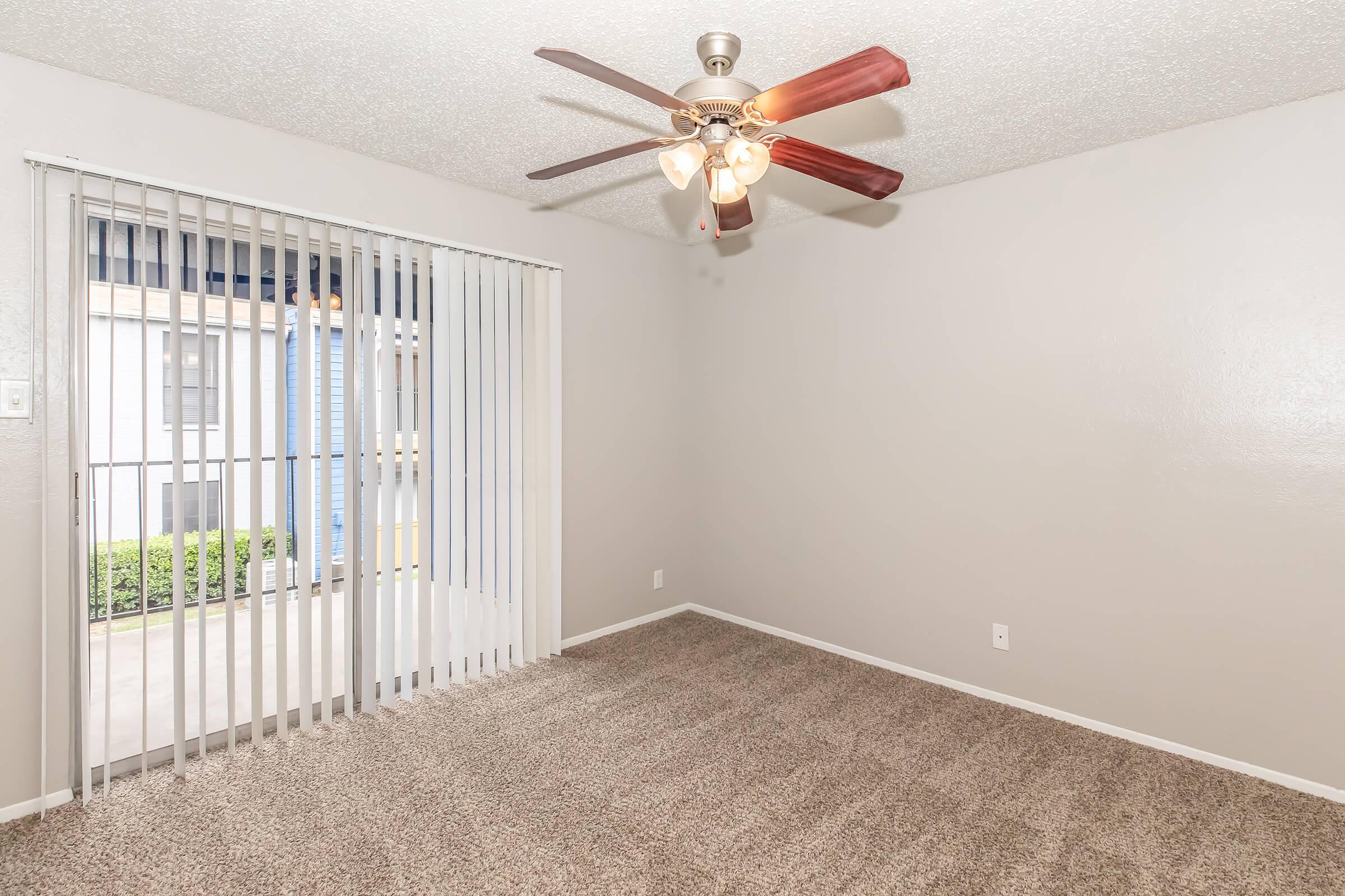A small, carpeted living room featuring a ceiling fan with wooden blades, light beige walls, and a sliding glass door with vertical blinds that leads outside. The room has a bright and airy feel, with natural light coming through the door and a view of greenery outside.