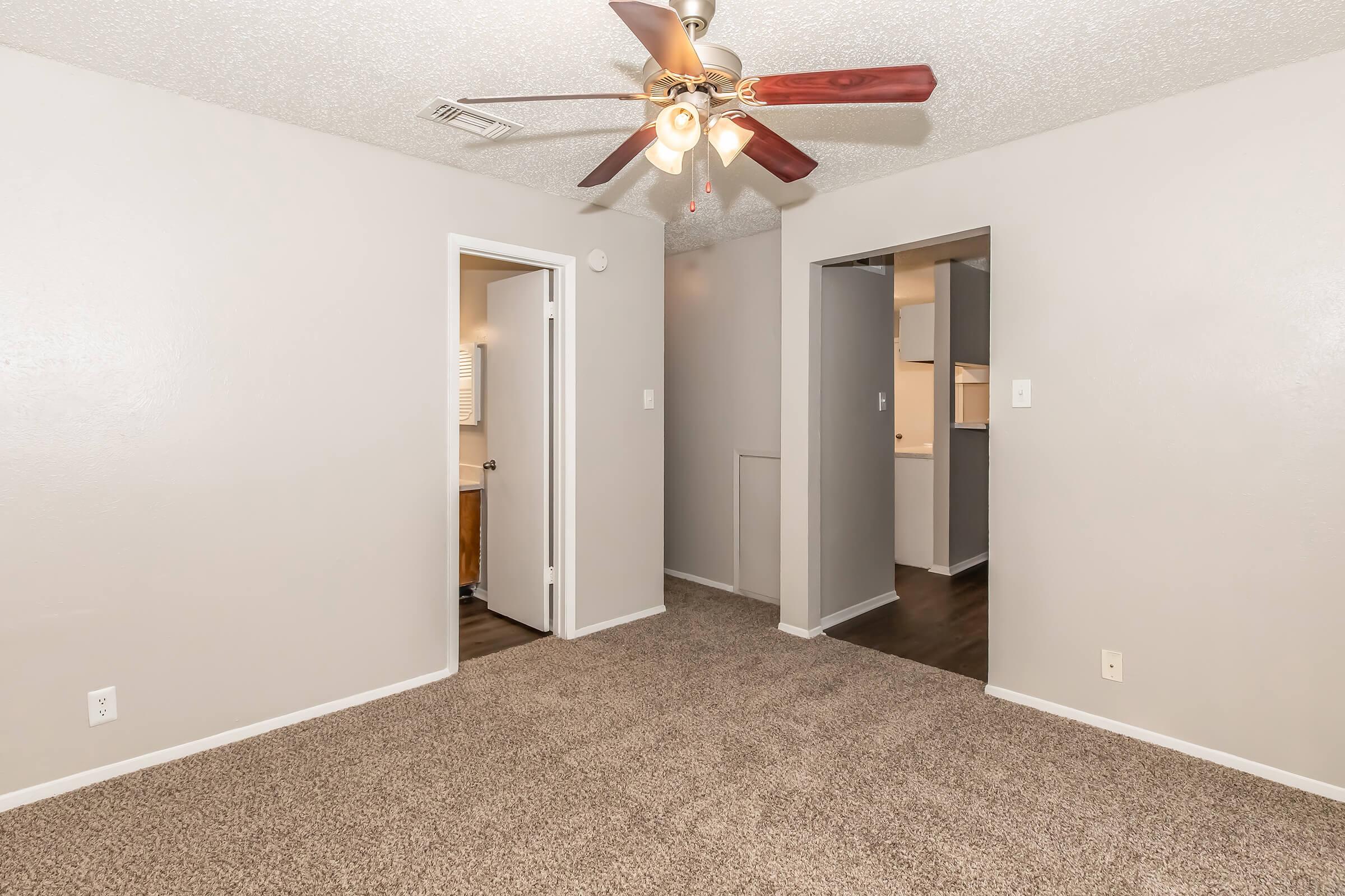 A spacious living area with light brown carpet, featuring a ceiling fan. Off to the side, there are two doorways leading to adjacent rooms. The walls are painted in a neutral tone, enhancing the open feel of the space.