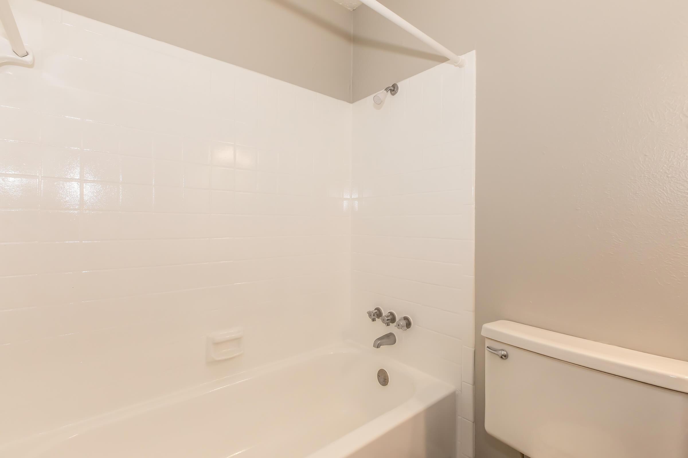 A clean and minimalist bathroom featuring a white bathtub with a shower curtain rod, a white tiled wall, and a modern toilet. The wall is painted in a light gray color, providing a neutral backdrop to the space.