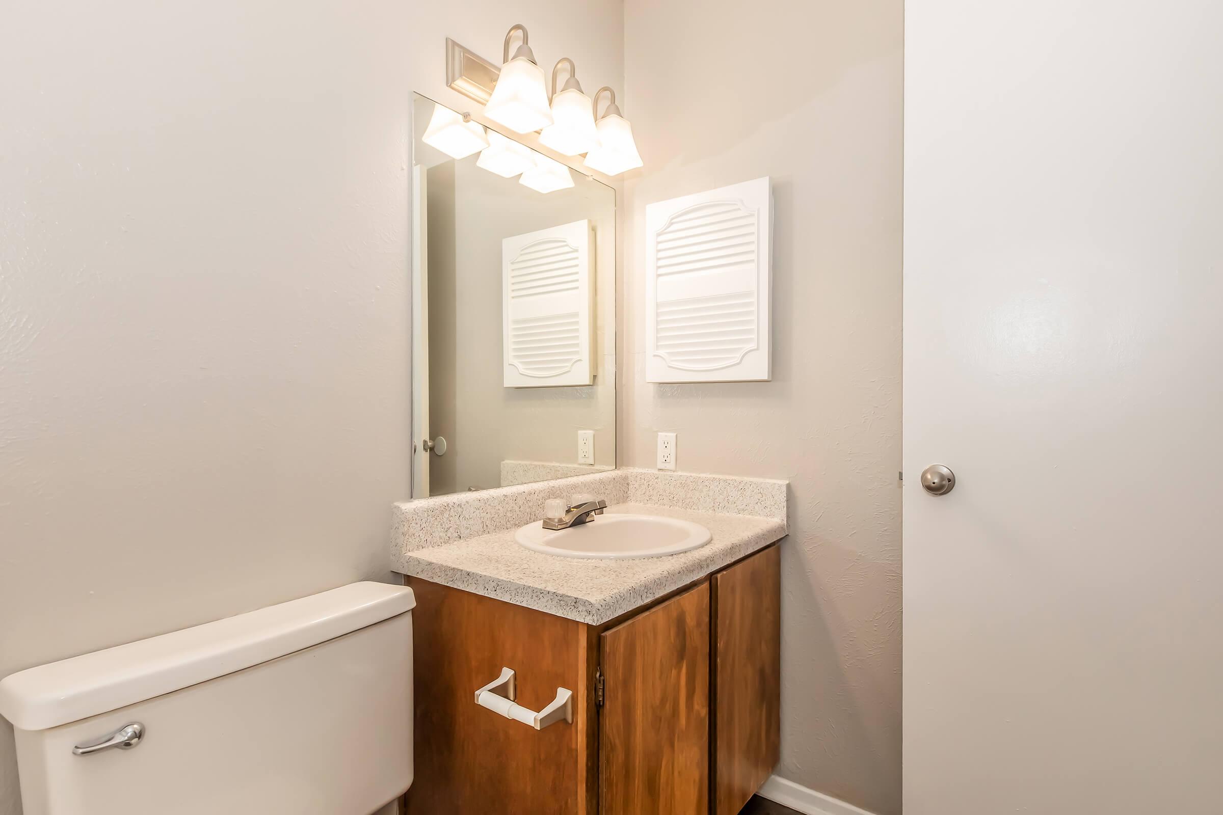 A small bathroom featuring a granite counter with a sink, a mirror above, and three light fixtures. There is a wooden cabinet beneath the sink, a white toilet, and a door partially closed on the right side. The walls are painted light gray, creating a clean and modern appearance.