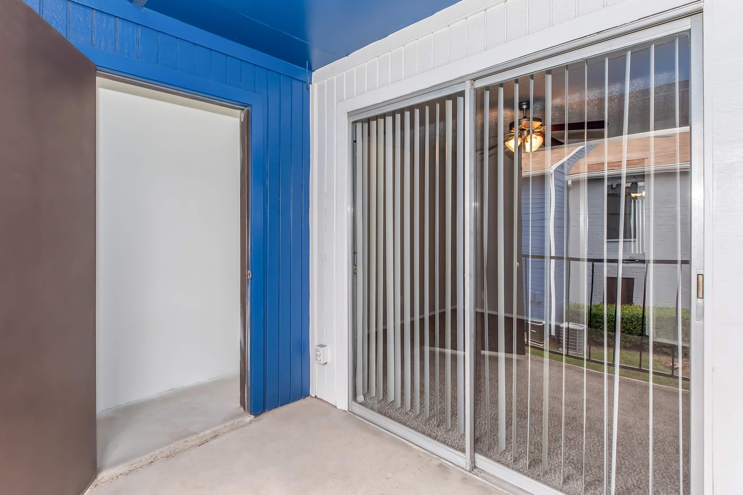 An entryway featuring a blue wall on the left, leading to a doorway that opens to a light-colored interior space. On the right, there are sliding glass doors with vertical blinds, providing a view of a small outdoor area with grass and neighboring buildings through the transparent panels.