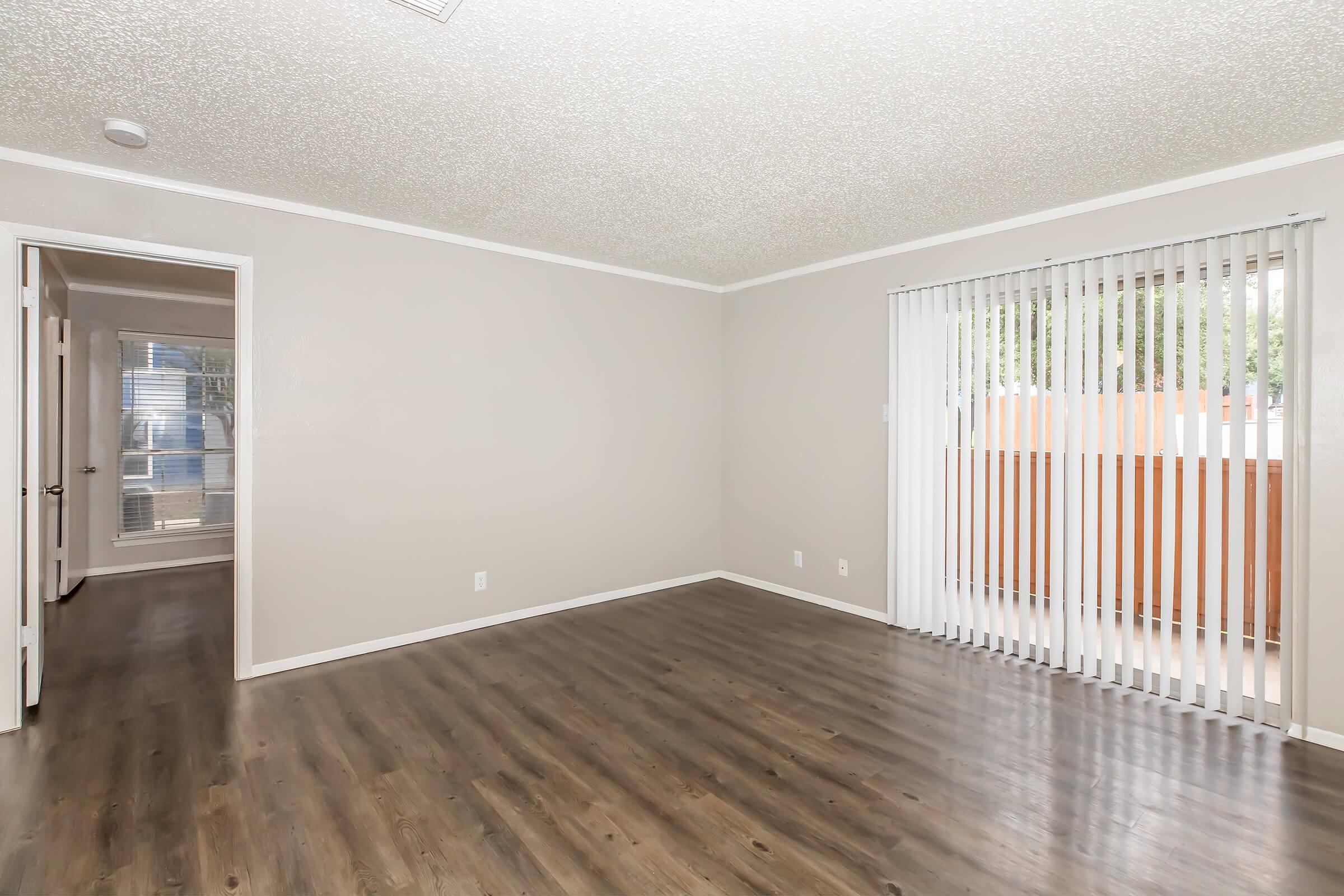 A spacious, empty room with light gray walls and a textured ceiling. The floor is dark hardwood. On one side, there is a sliding glass door with vertical blinds, providing access to an outdoor area. A doorway leads to another room, suggesting an open layout. The space is bright and inviting.