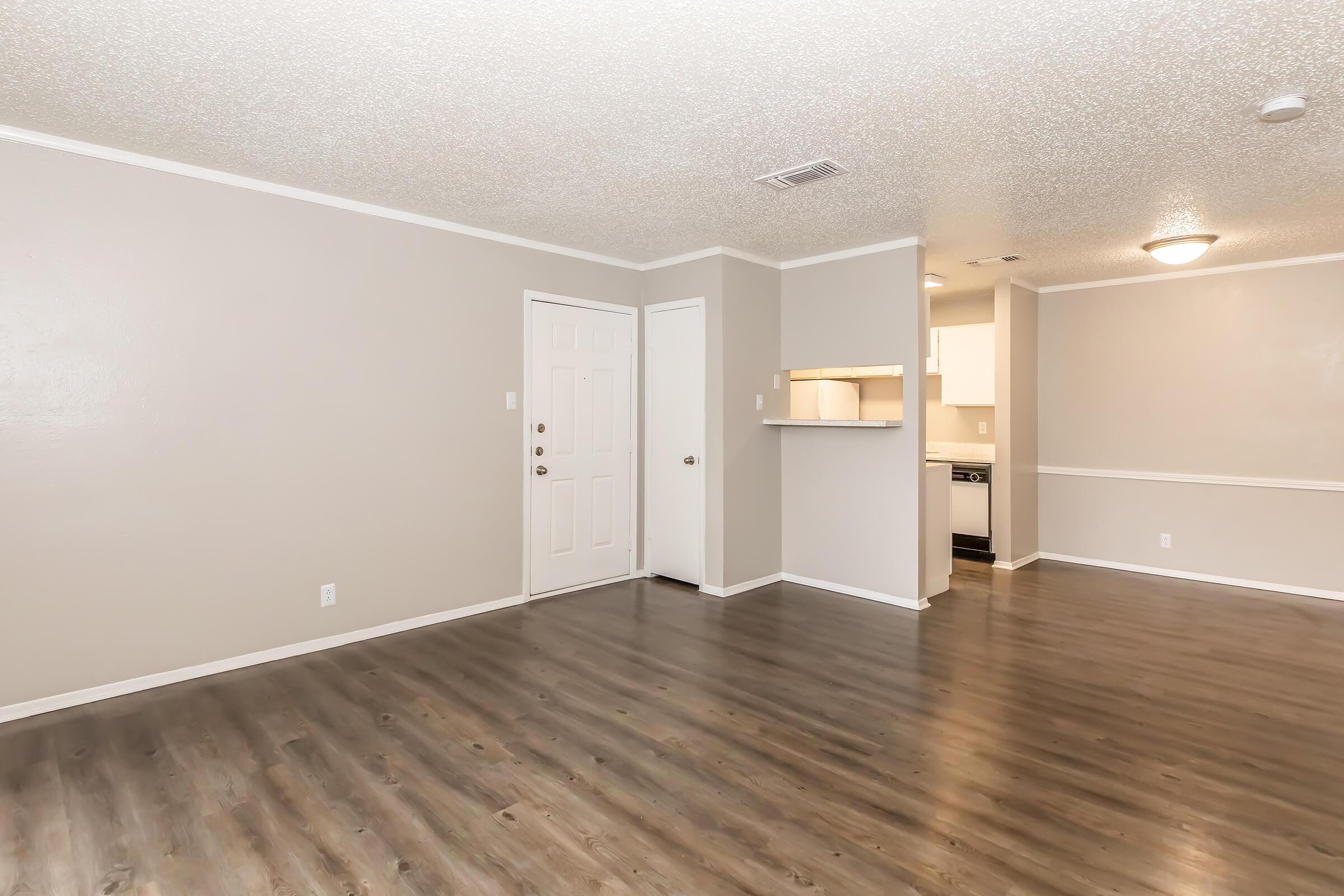 A spacious, empty living room with light gray walls and new laminate flooring. The room features a front door, an open kitchen area with white cabinets visible in the background, and a small bar counter. Natural light brightens the space, creating a welcoming atmosphere.