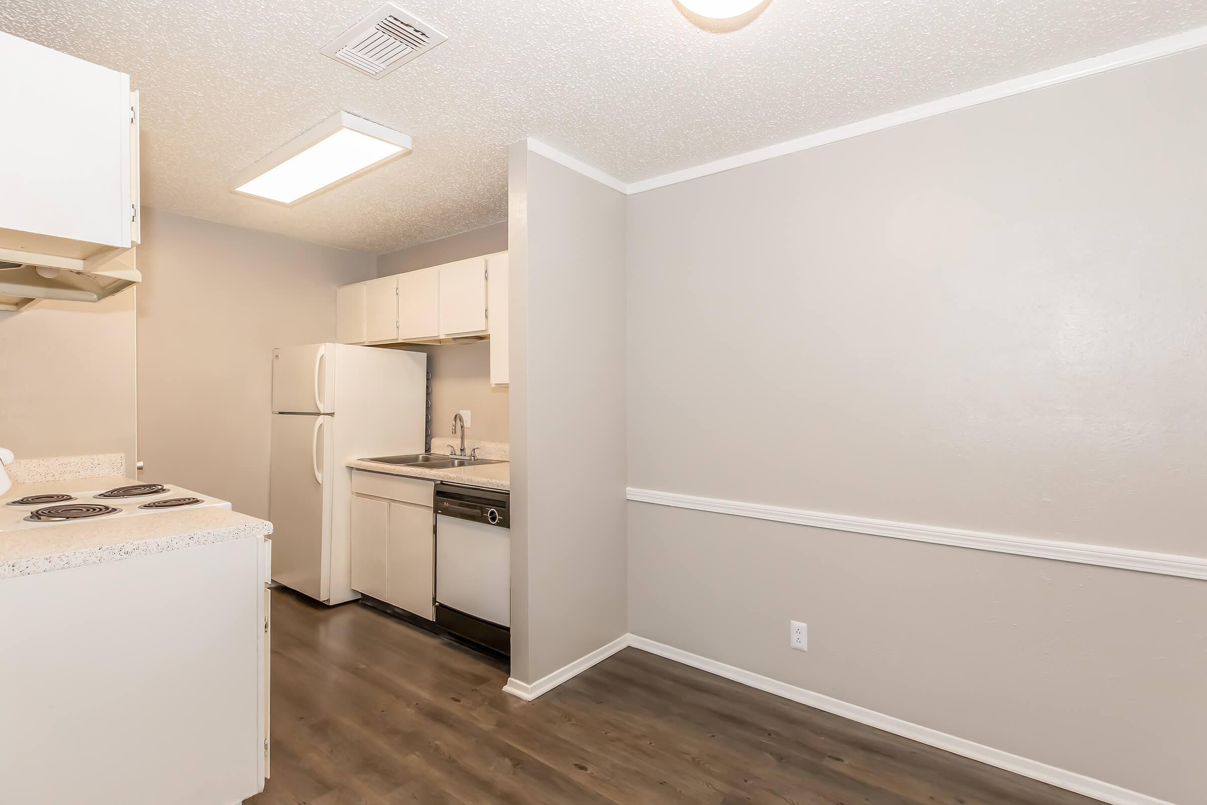 A modern kitchen featuring white cabinets, a stainless steel sink, a dishwasher, and a refrigerator. The space has a neutral color scheme with light gray walls and dark flooring. A bright overhead light fixture illuminates the area, creating a clean and open look.