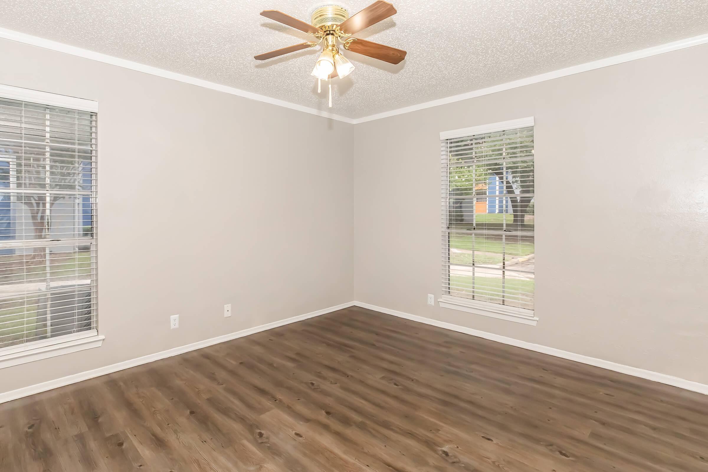 A spacious room featuring light brown walls, a ceiling fan with wooden blades, and large windows with white blinds. The flooring is a dark wood laminate, and it appears empty, creating a bright and airy atmosphere with natural light coming through the windows.
