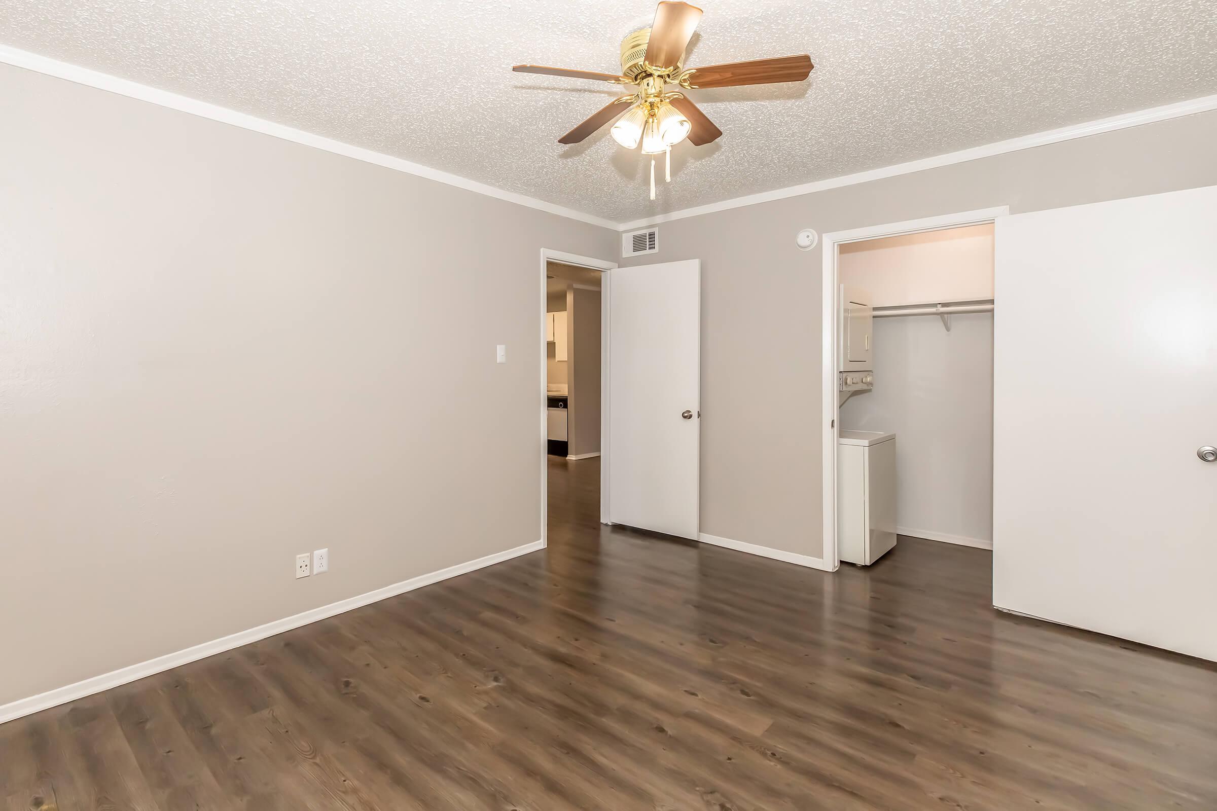 A well-lit, empty room with light gray walls and brown laminate flooring. It features a ceiling fan with wooden blades, two doors leading to adjacent rooms, and a closet area. A small laundry space is visible through one of the doors. The overall appearance is clean and modern.