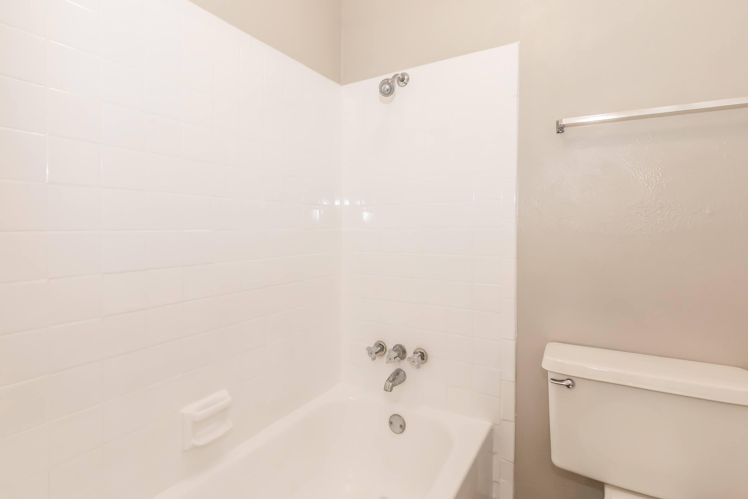 A clean and simple bathroom featuring a white tiled bathtub with chrome faucets and a showerhead. A white toilet is located on the side, and there is a towel bar mounted on the wall. The overall color scheme is neutral with light walls and bright tiles, creating an airy atmosphere.