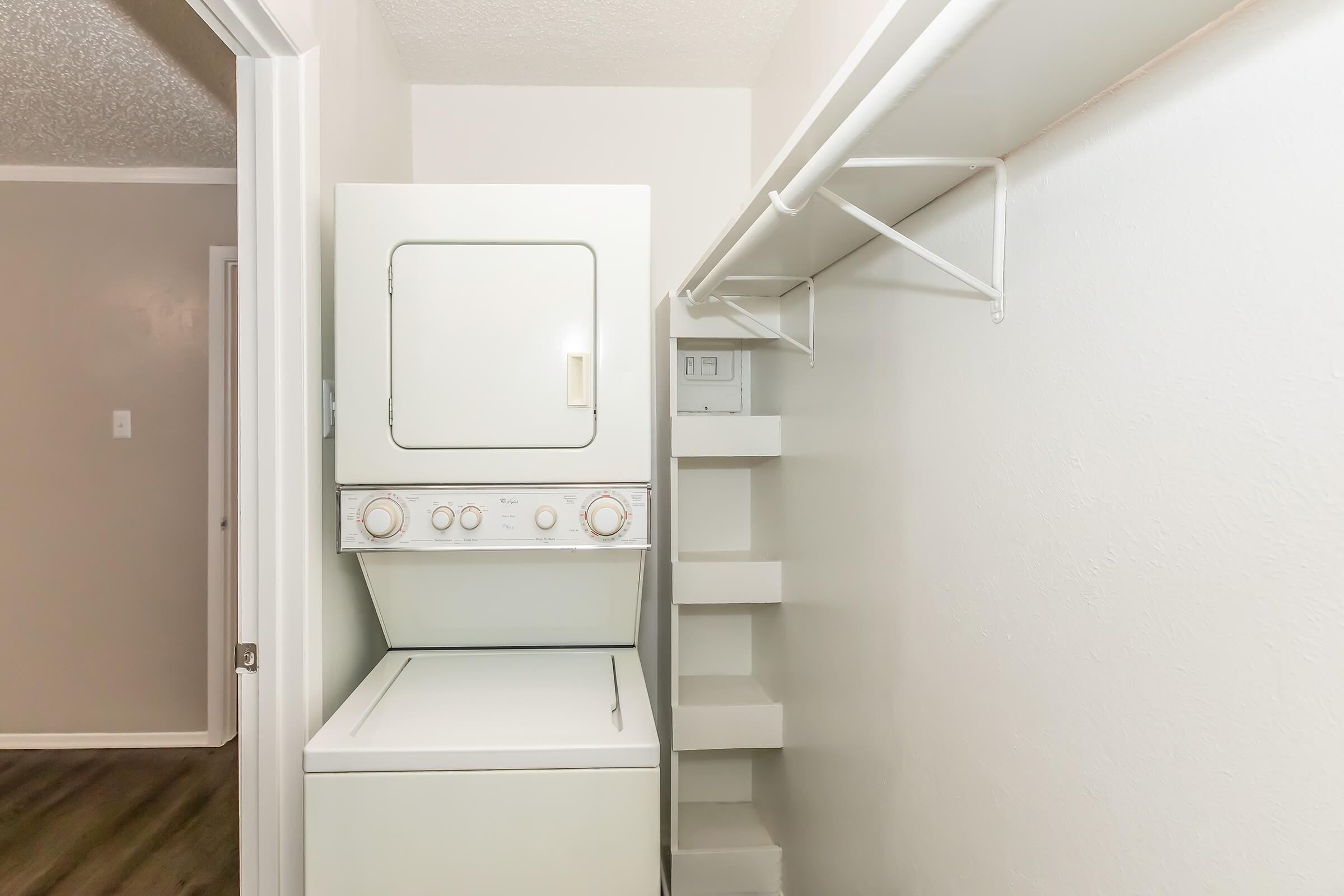 A compact laundry area featuring a stacked washing machine and dryer unit beside a set of open shelves. The walls are painted light colors, and there is a door leading to another room in the background, showcasing a clean and organized space.