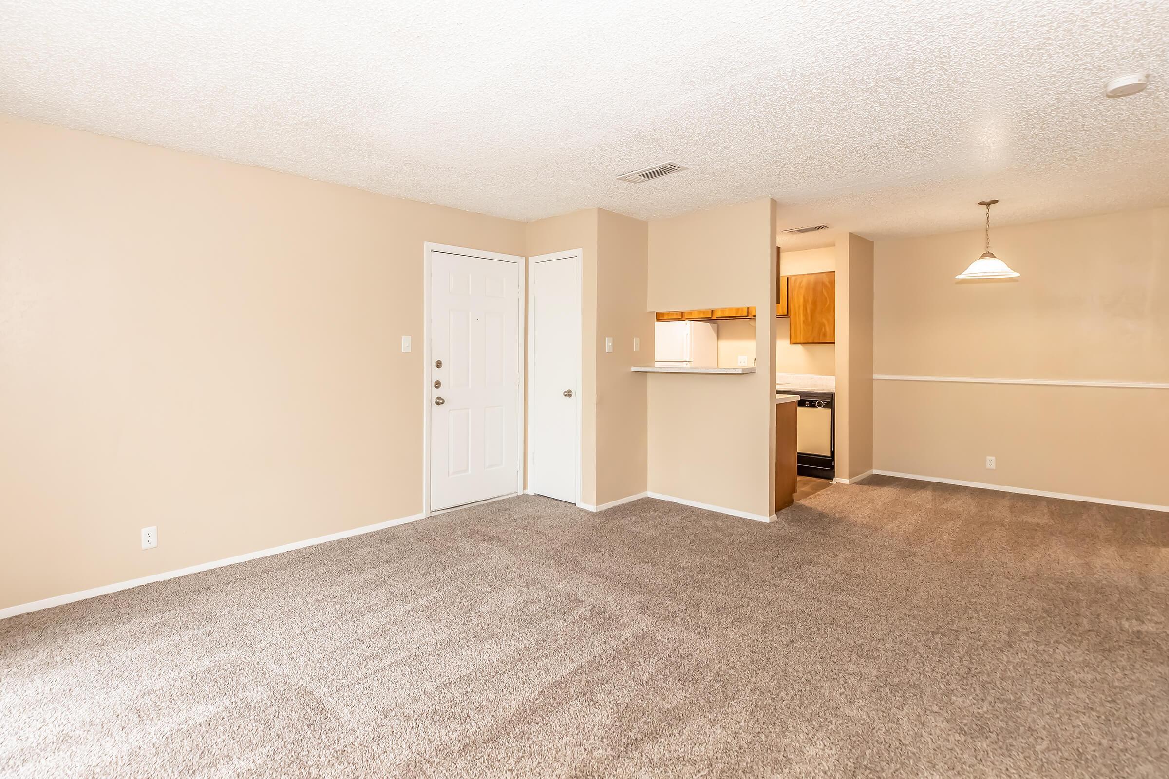 Spacious living area with beige walls and plush carpeting, featuring a front door, an open kitchen with wooden cabinets in the background, and a pendant light fixture above. The layout is bright and inviting, perfect for comfortable living.