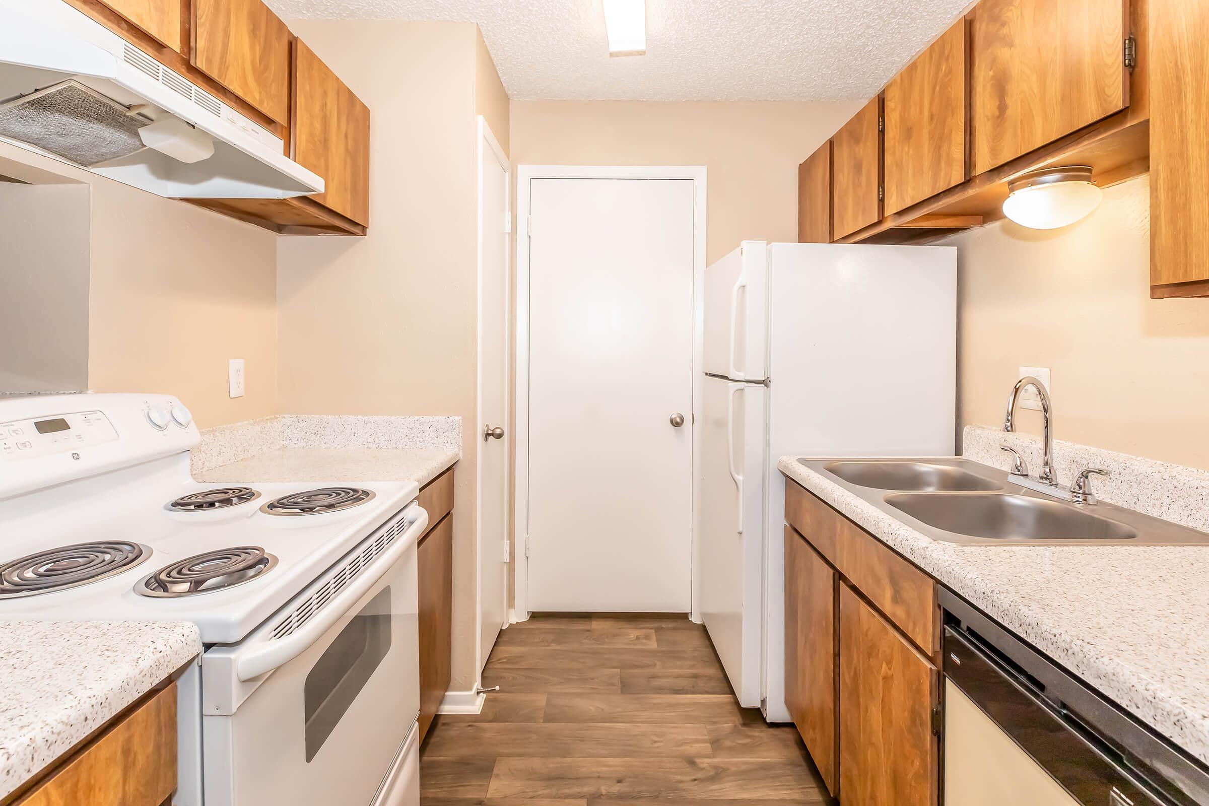 A well-lit kitchen featuring wooden cabinets, a white stove, and a refrigerator. The countertop is made of light-colored granite, complemented by a double sink. The walls are painted in a warm, neutral tone, and the floor is made of attractive wood-like laminate. A door leads to another room.