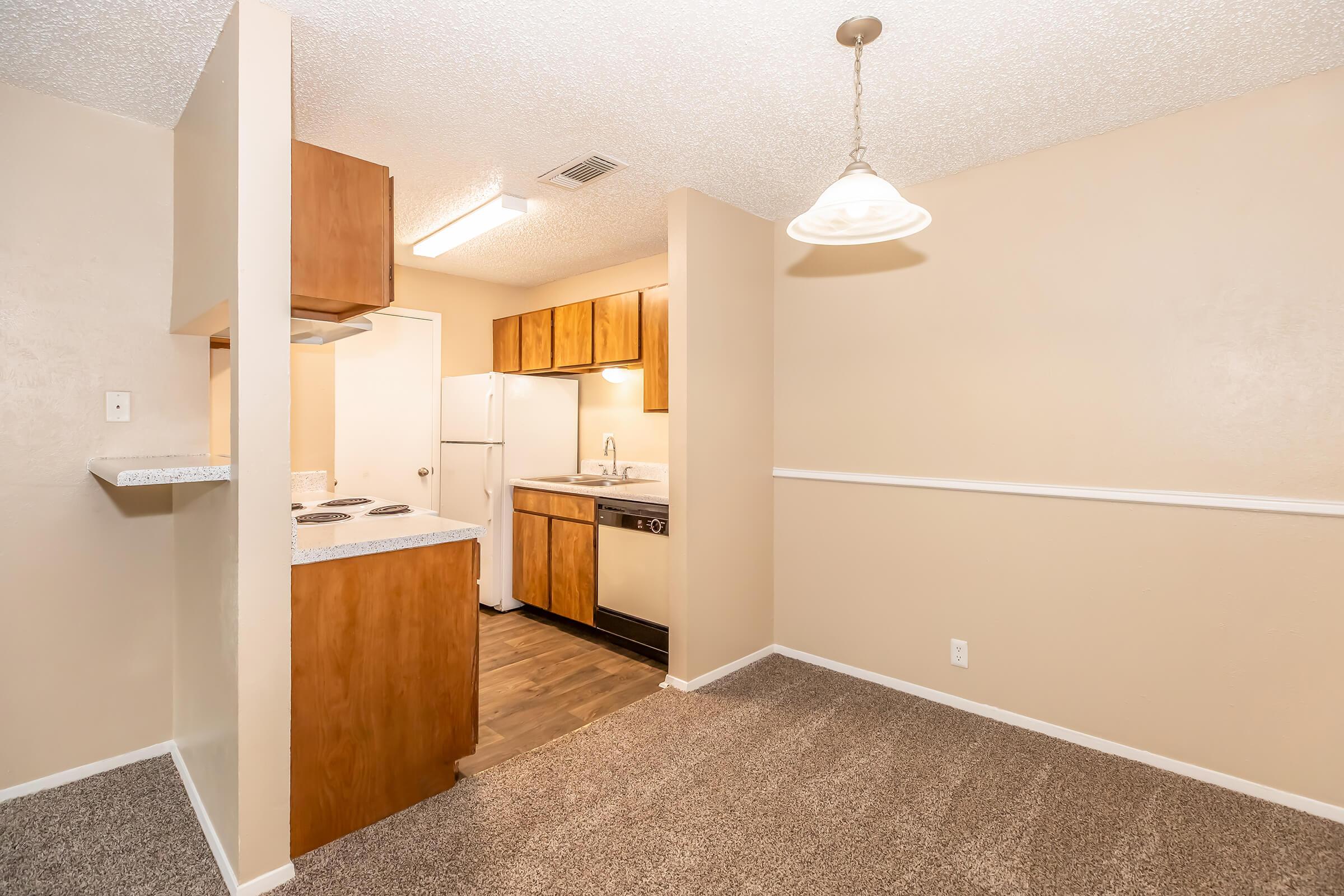 A small, cozy kitchen with wooden cabinets and modern appliances, including a stove, refrigerator, and dishwasher. The room features a textured ceiling with a light fixture and a warm beige wall color. A portion of the kitchen includes a bar area with countertop space. The floor is covered in light-colored carpet.