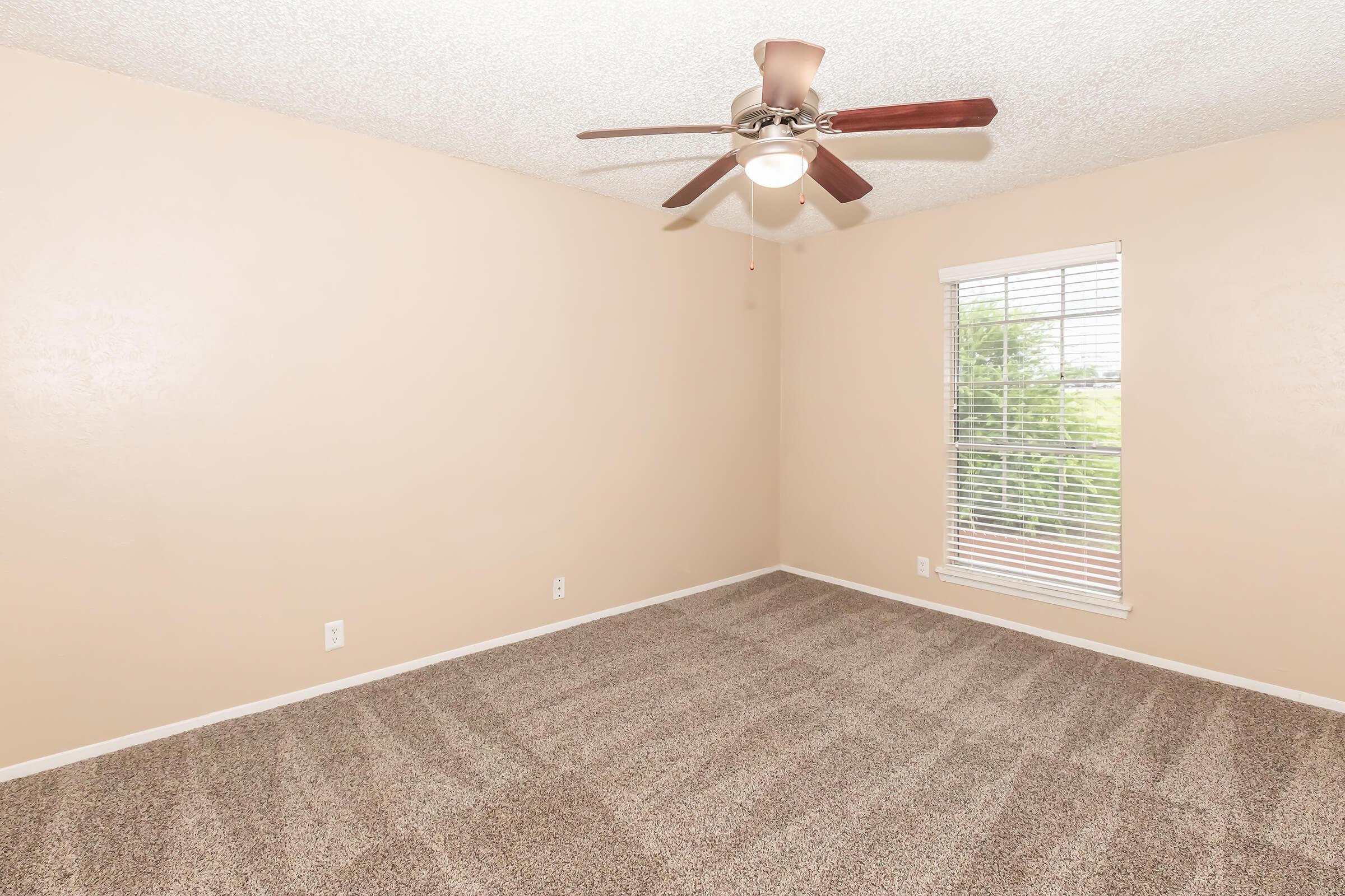 A spacious, empty room with light beige walls and brown carpet. A ceiling fan with wooden blades hangs from the ceiling. There are white window blinds on a window allowing natural light, providing a view of greenery outside. The room is well-lit and ready for furnishings.
