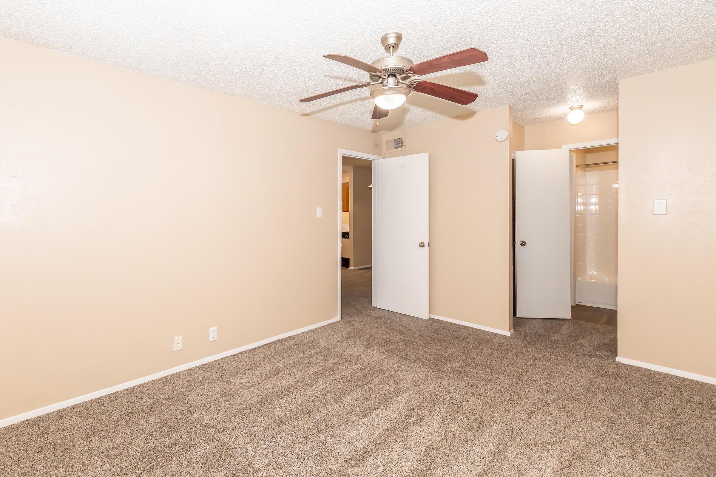 Interior view of a spacious room featuring beige walls, carpet flooring, and a ceiling fan with wooden blades. There are two doors leading to adjacent rooms, and a bathroom doorway on the right. The room is well-lit and inviting, suitable for various uses.