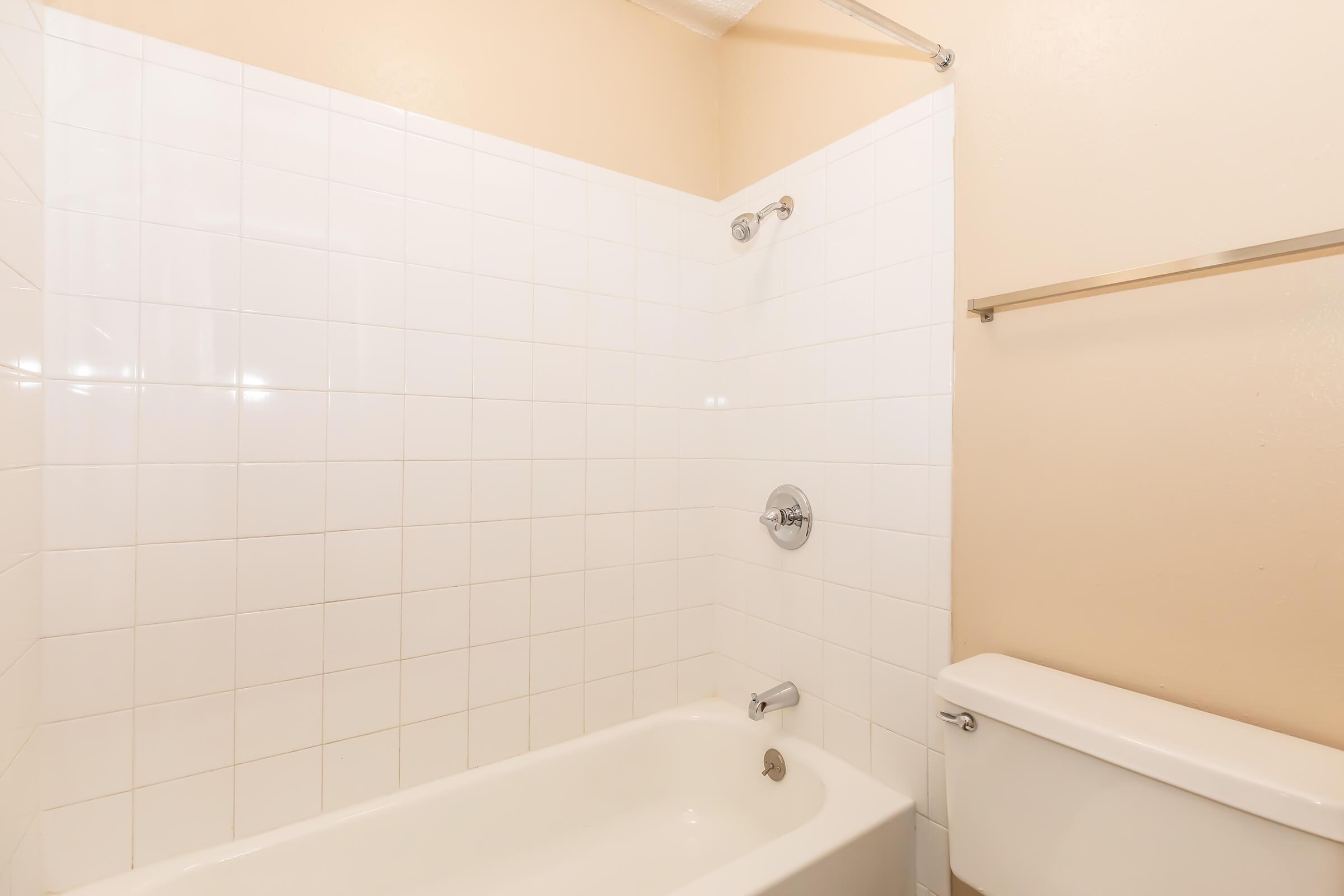 A clean, simple bathroom with white tiled walls. There is a bathtub with a showerhead above it and a chrome faucet. Next to the tub, a standard toilet is visible. The walls are painted a light beige color, creating a bright and airy atmosphere. The overall appearance is neat and minimalistic.