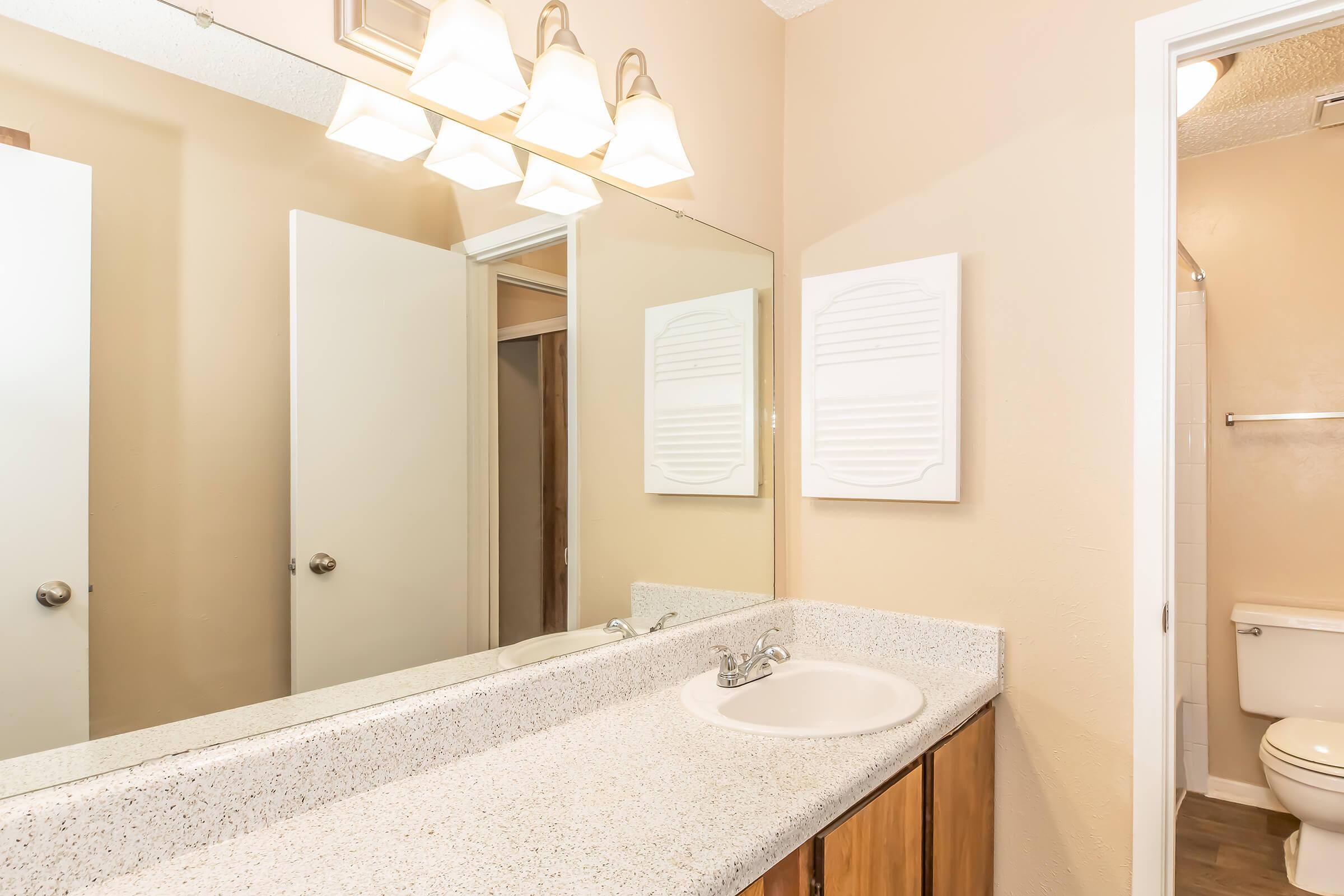 A well-lit bathroom featuring a double sink vanity with a speckled countertop, a large mirror above, and three light fixtures. Beige walls complement the wooden cabinetry below the sink. A towel rack is mounted on the wall, with a door leading to a shower area and another door to a toilet room.