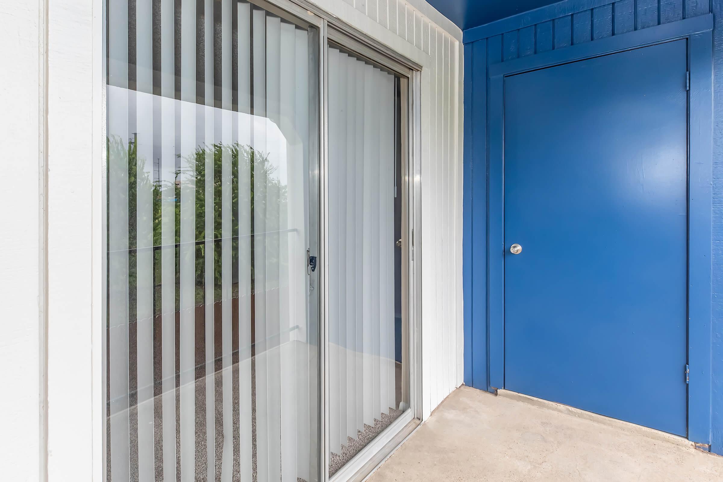 A porch area featuring a sliding glass door with vertical blinds beside a blue door. The surface is concrete, and there are green plants visible outside through the glass. The walls are painted white and blue, creating a modern entrance space.