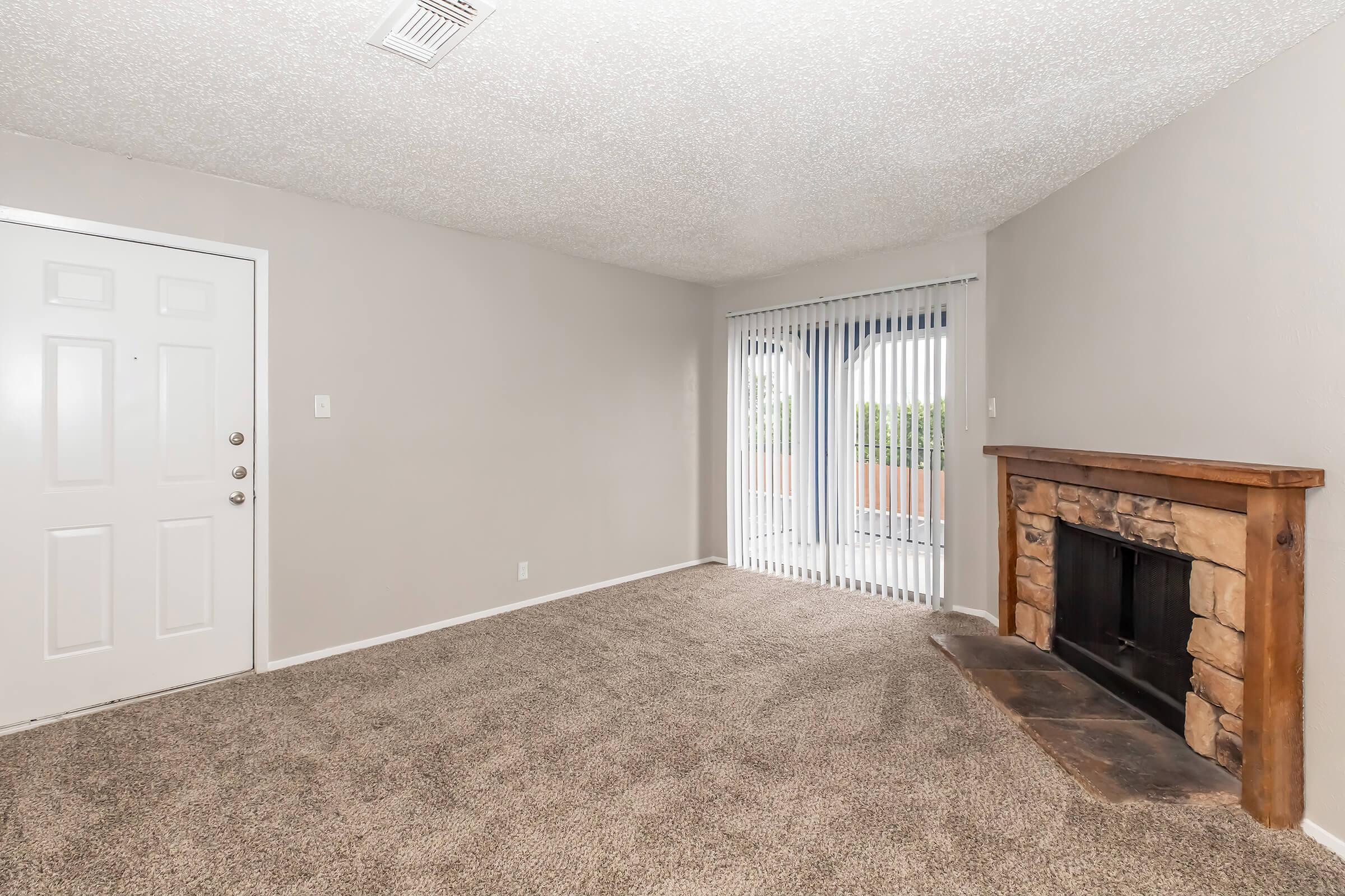 A spacious living room featuring beige walls and carpet, with a decorative stone fireplace on one side. There’s a glass sliding door with vertical blinds leading outside, and a front door visible on the left side of the image. Natural light is coming through the sliding door, enhancing the room's brightness.