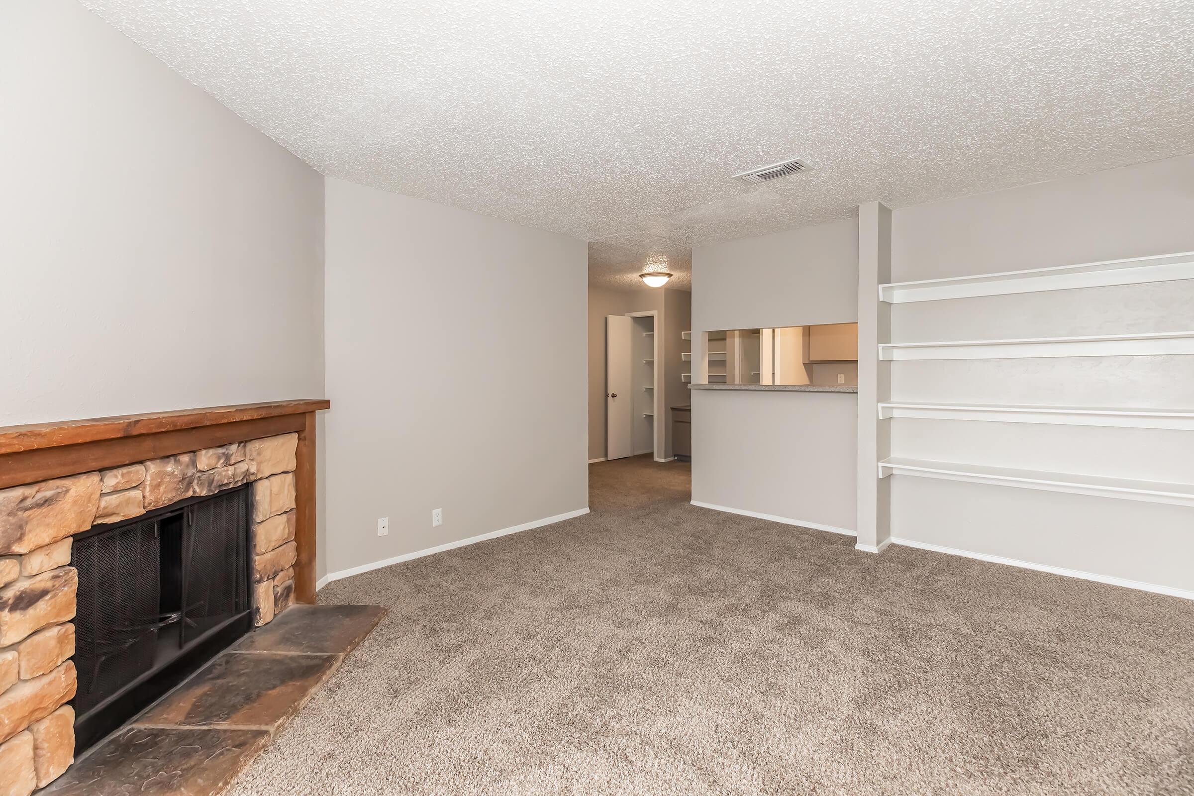 A cozy living room featuring a stone fireplace, soft carpet flooring, and light-gray walls. Built-in shelving is positioned near the fireplace, and a doorway leads to another room. The space is well-lit with an overhead light fixture, creating a warm and inviting atmosphere.