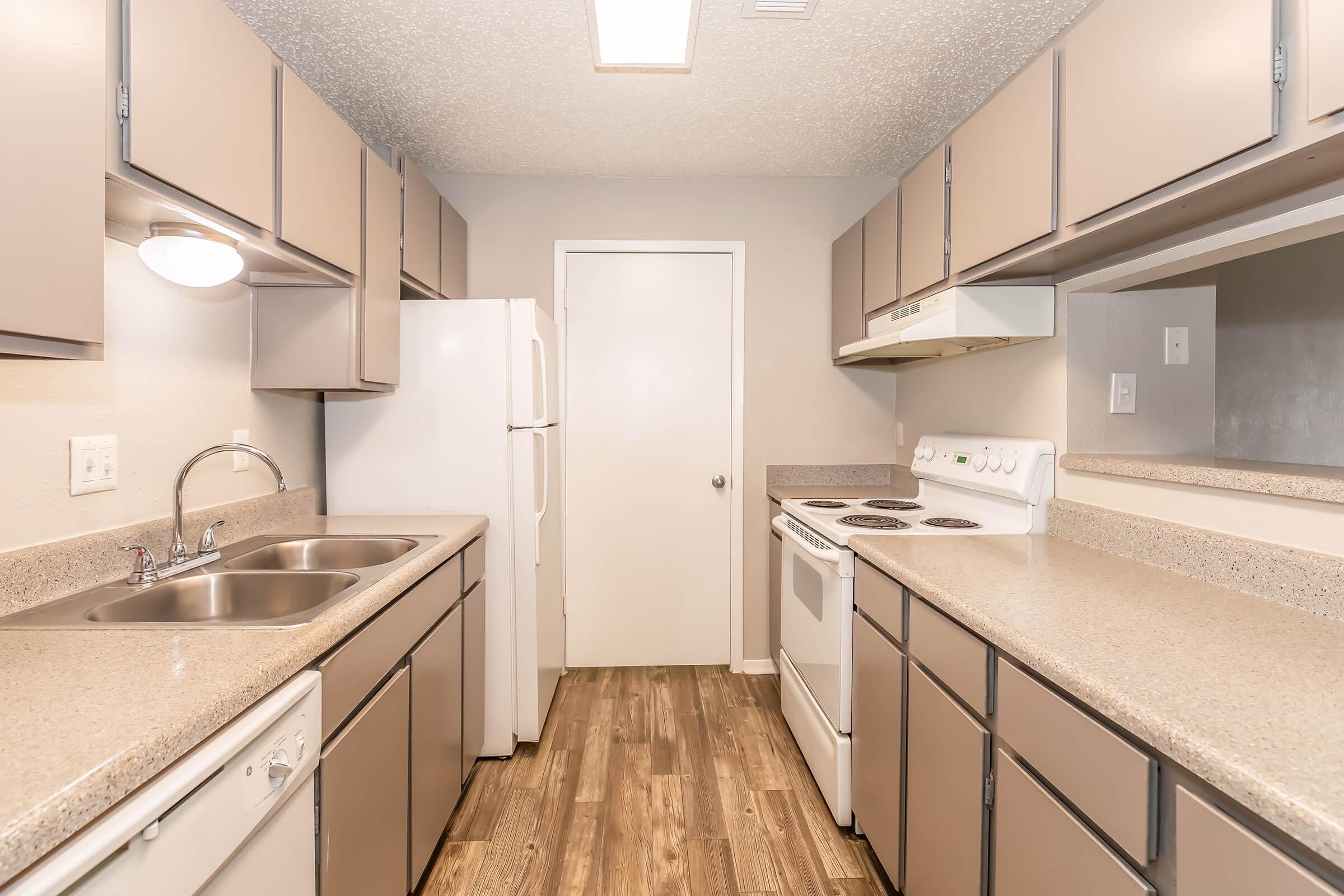 A modern kitchen featuring beige cabinets, stainless steel sink, white appliances including a refrigerator and stove, and a countertop with a textured surface. The flooring is wood-like, and the room is well-lit with overhead lighting. A door is visible at the end of the kitchen.