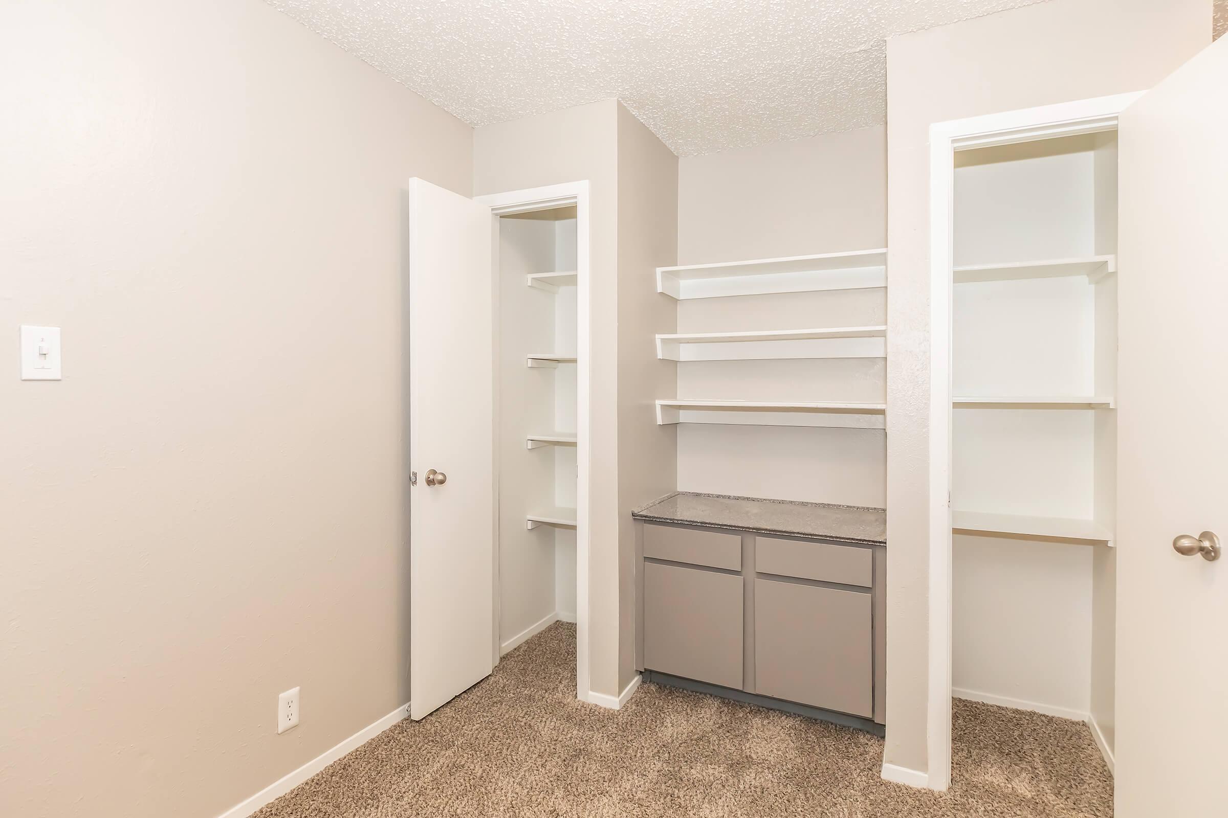 A well-lit, empty closet with beige walls and carpeting. The space features two open doors leading into separate sections with shelving, and a central cabinet with a dark countertop. The overall design is clean and modern.