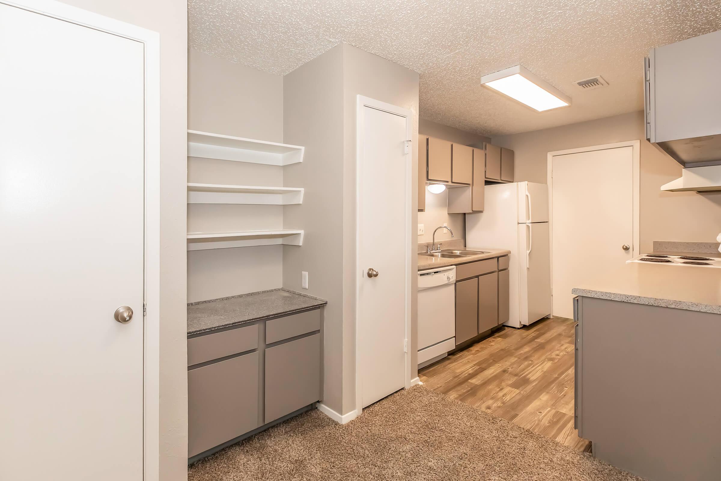 Modern kitchen interior featuring gray cabinets, a white refrigerator, and a dishwasher. The space includes a door leading to another room, open shelving, and a countertop. The floor is covered with a wood-like laminate, and the walls are painted in light tones, creating a bright and inviting atmosphere.